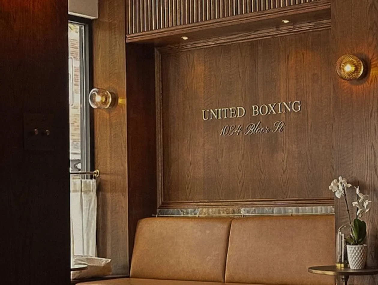 Interior of a room with wooden wall paneling, a sign that reads 'United Boxing 1034 Bloc Ct,' a brown couch, a small round table with a potted orchid plant, and window with natural light.