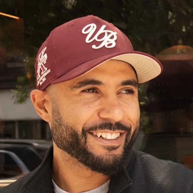 A smiling man with a beard wearing a maroon baseball cap with white lettering and a black jacket.