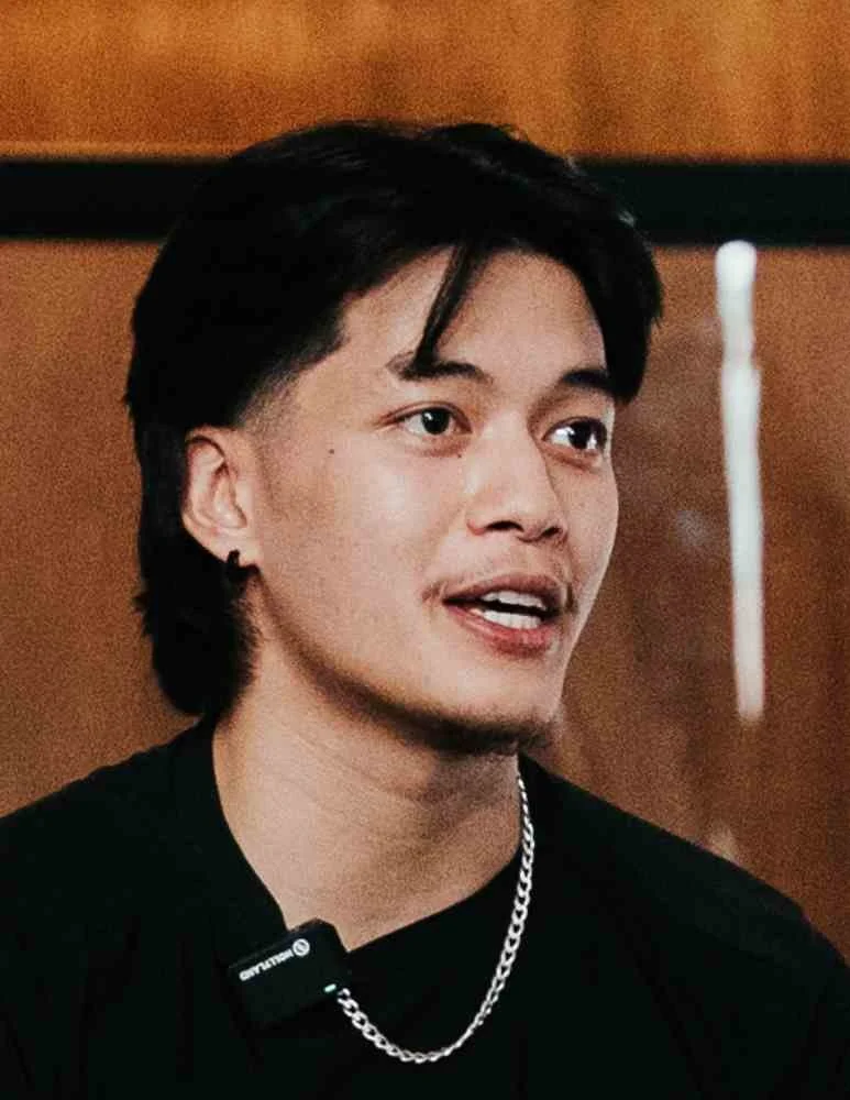 A young man with black hair, wearing a black shirt and silver chain, speaking in front of a wooden background.