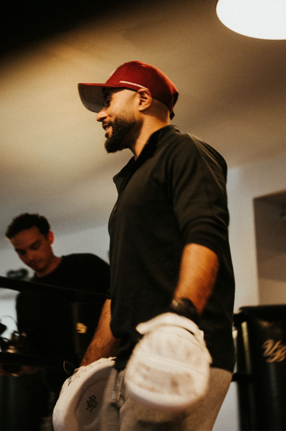 A man wearing a red cap, black shirt, and white shoes, standing indoors near a piano with another person in the background.