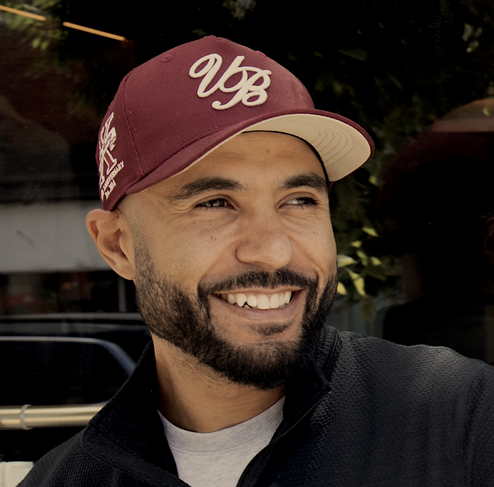 Smiling man with a beard wearing a maroon and white baseball cap and a black jacket.