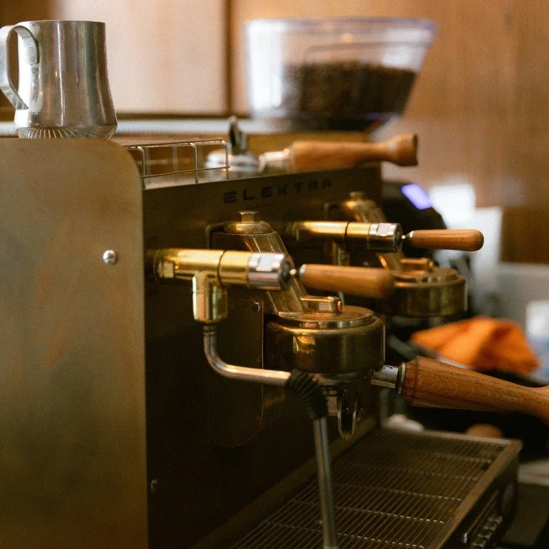 Close-up of an espresso machine with wooden handles and a small metal pitcher on top, with coffee beans in a transparent container in the background.