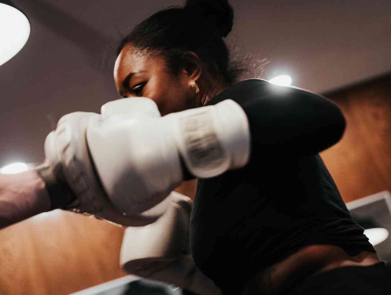 A woman practicing boxing, wearing boxing gloves and a black top, in a gym or training room.