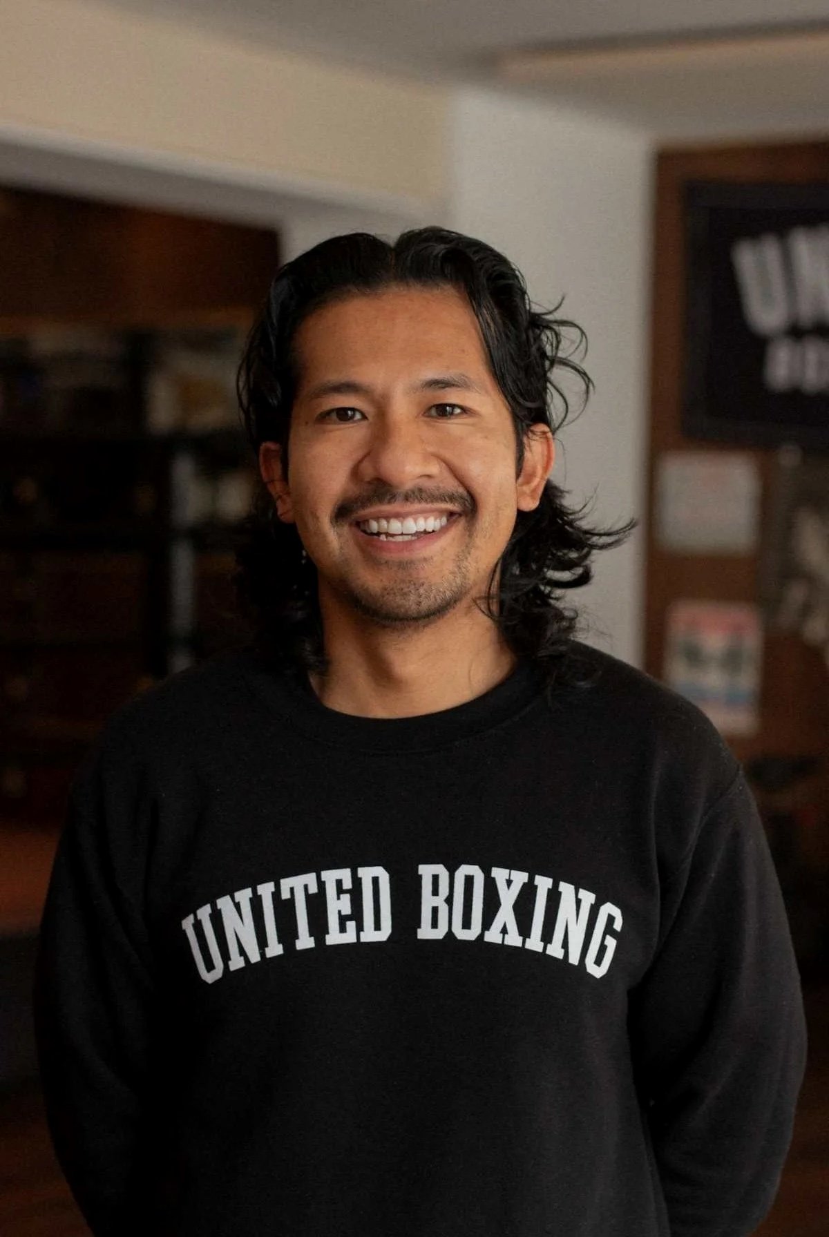 A man smiling, wearing a black sweatshirt with the words 'United Boxing' on it, standing indoors in a room with wooden furniture and a bulletin board in the background.