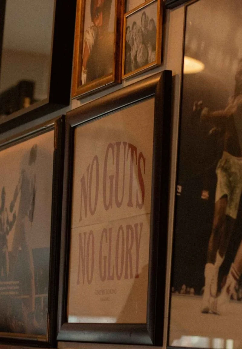 Close-up of a framed poster with the text "NO GUTS, NO GLORY" surrounded by other framed pictures on a wall.
