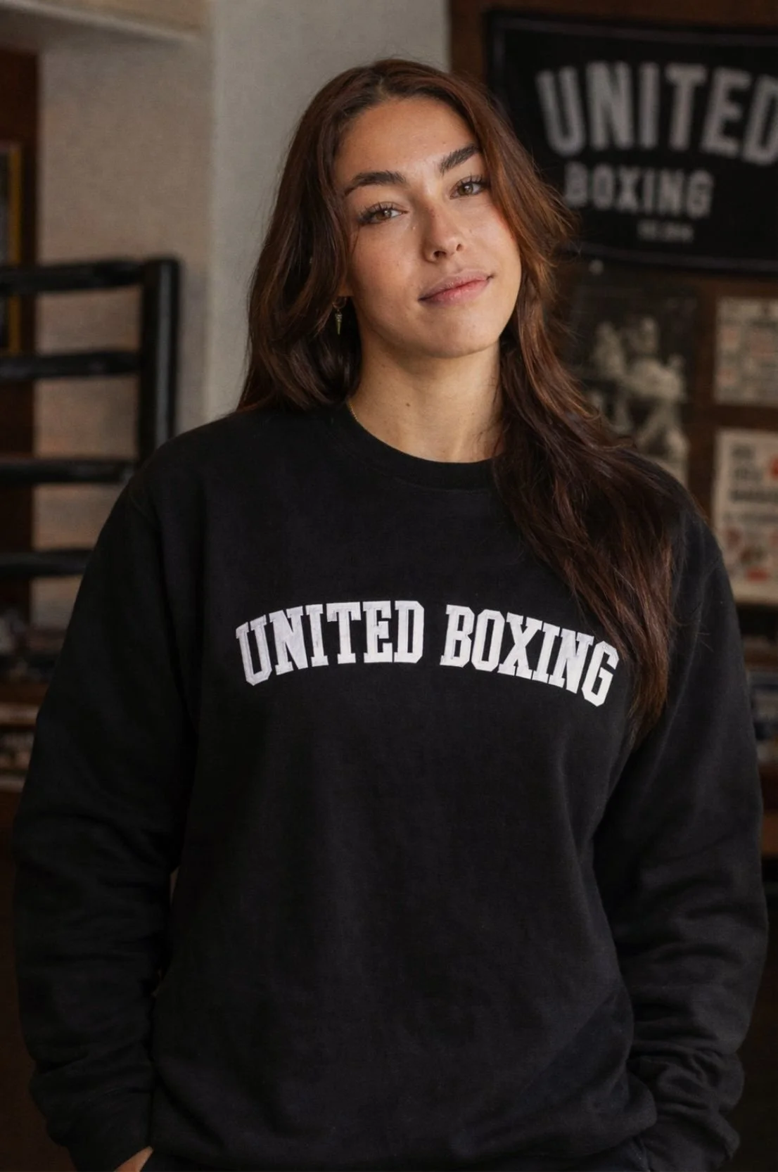 Young woman with brown hair wearing a black sweatshirt with "United Boxing" written on it, standing indoors with a neutral expression.