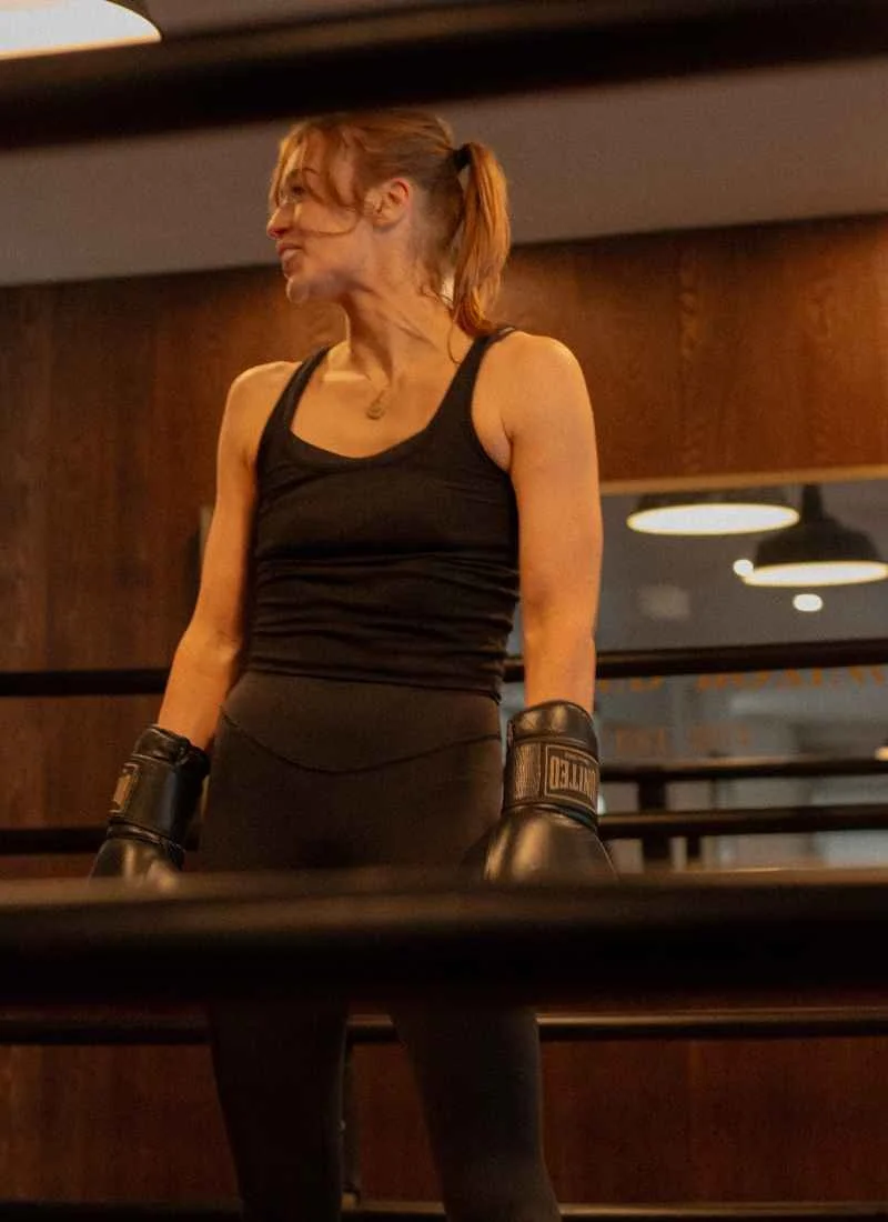 A woman in black athletic attire and boxing gloves, standing in a gym with wooden walls and ceiling lights.