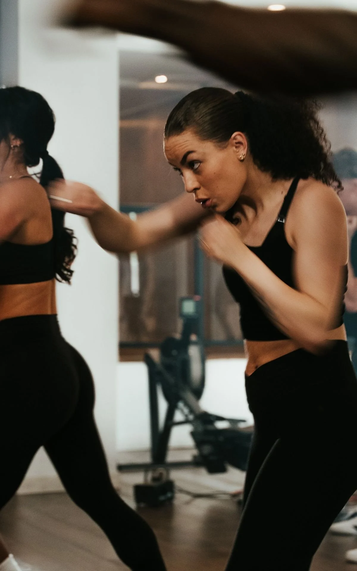 Women practicing boxing in a gym, wearing black sports bras and leggings, with one woman throwing a punch at her training partner.