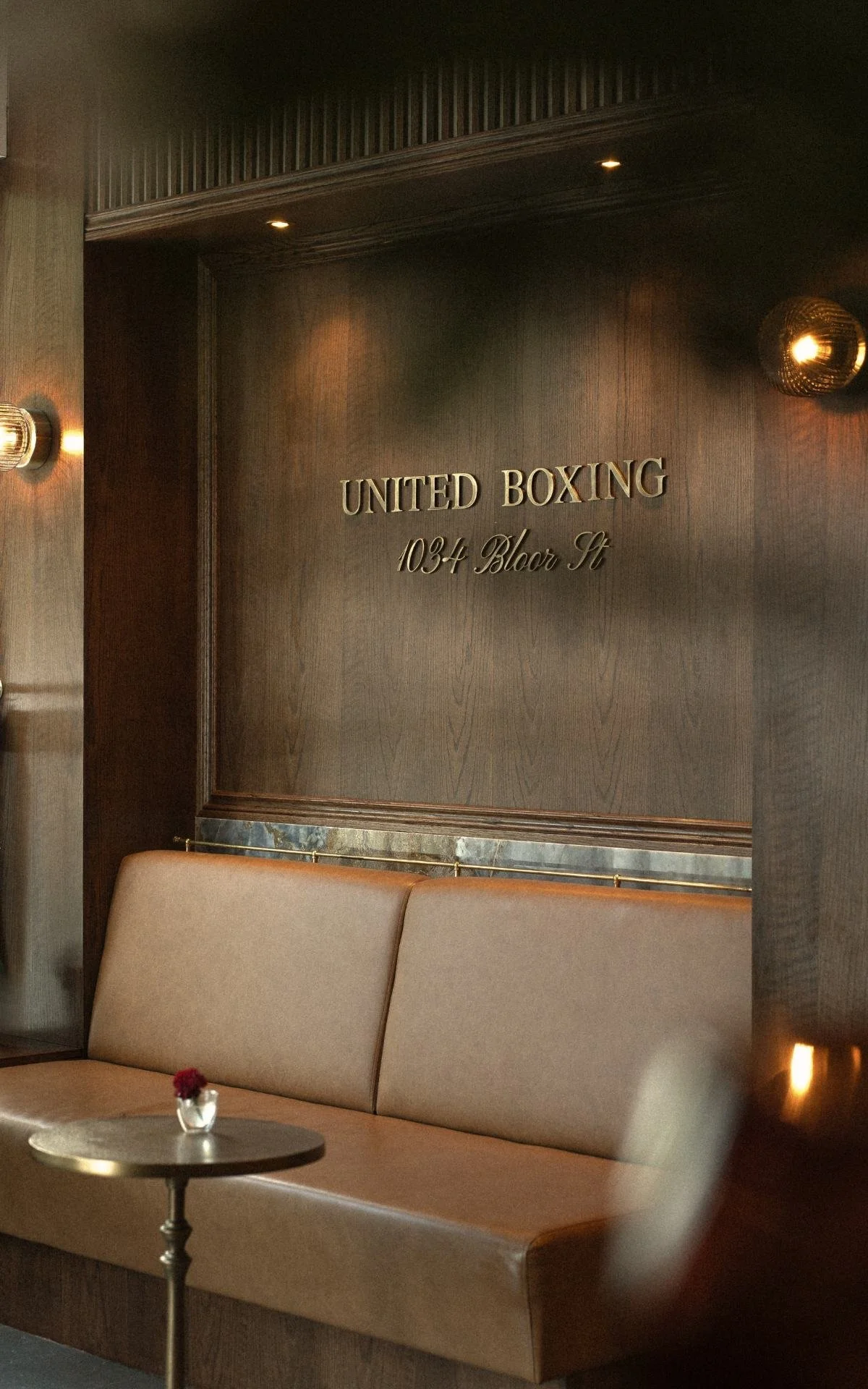 Interior of a boxing gym lounge area with a beige bench, a small round table with a small flower vase, dark wooden walls, and a sign reading 'United Boxing 1034 Bloor St'.