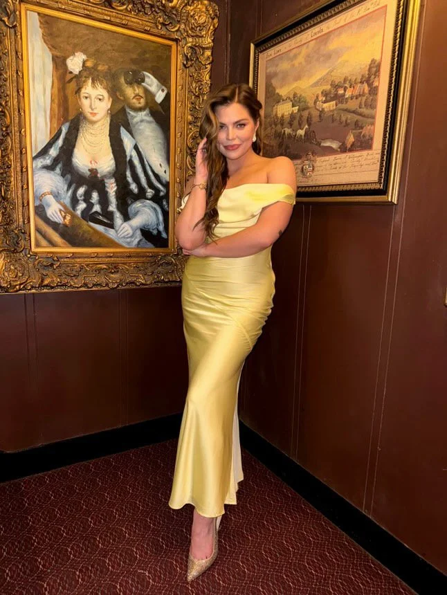 A woman in a yellow off-the-shoulder gown poses in front of two framed paintings in an indoor setting. One painting depicts a woman and a man with dark hair, and the other shows a scenic landscape.