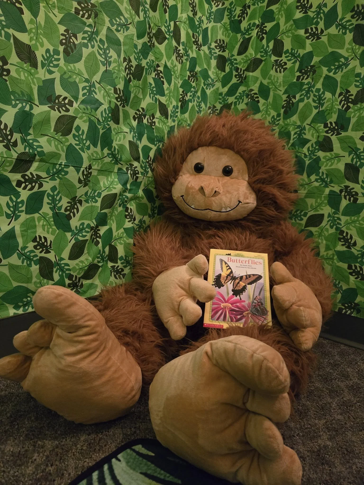 A large plush Sasquatch, sitting against a green leafy patterned background, holding a children's book titled 'Butterflies'.