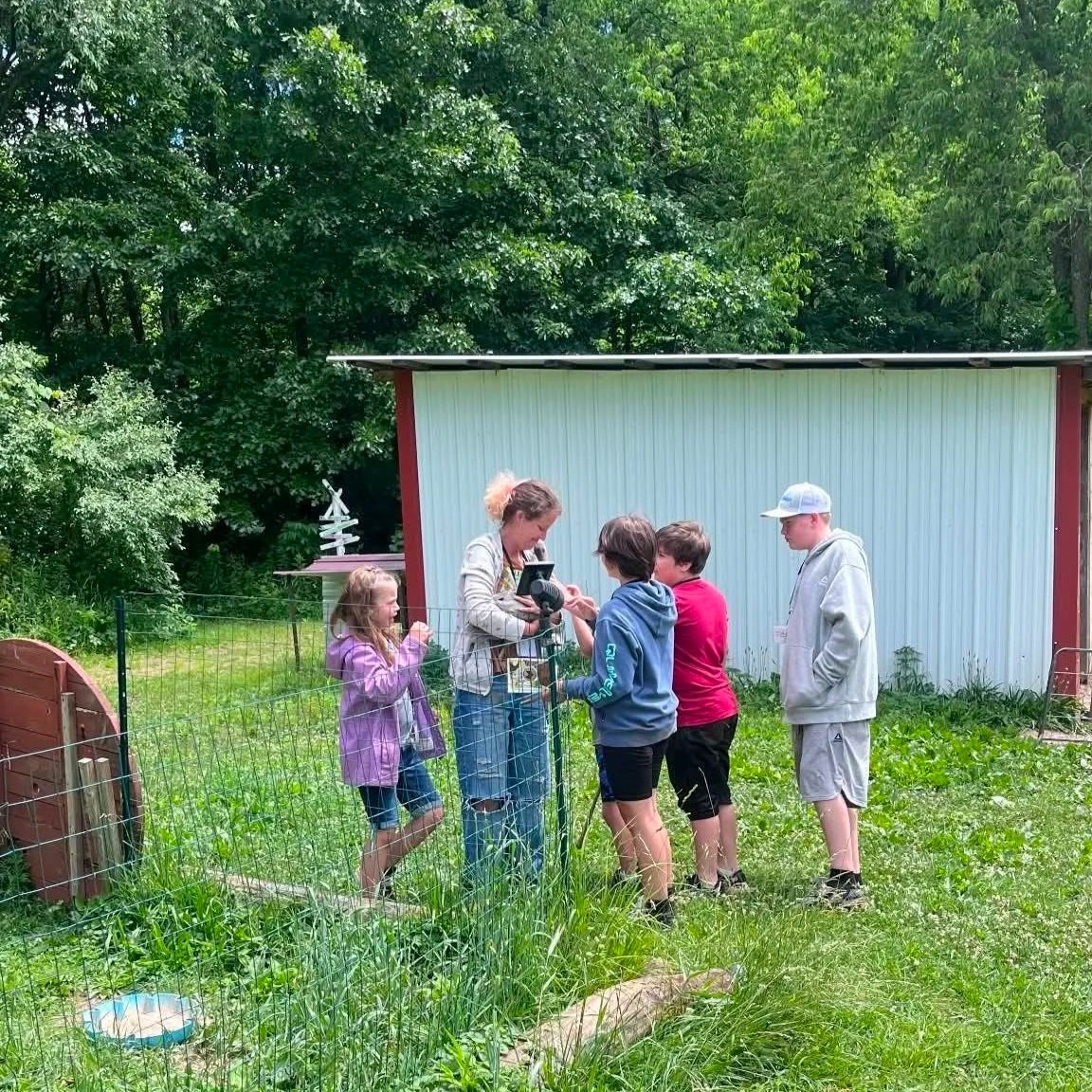 A group of children gathered around a woman holding a duck in a backyard, with a shed and trees in the background.