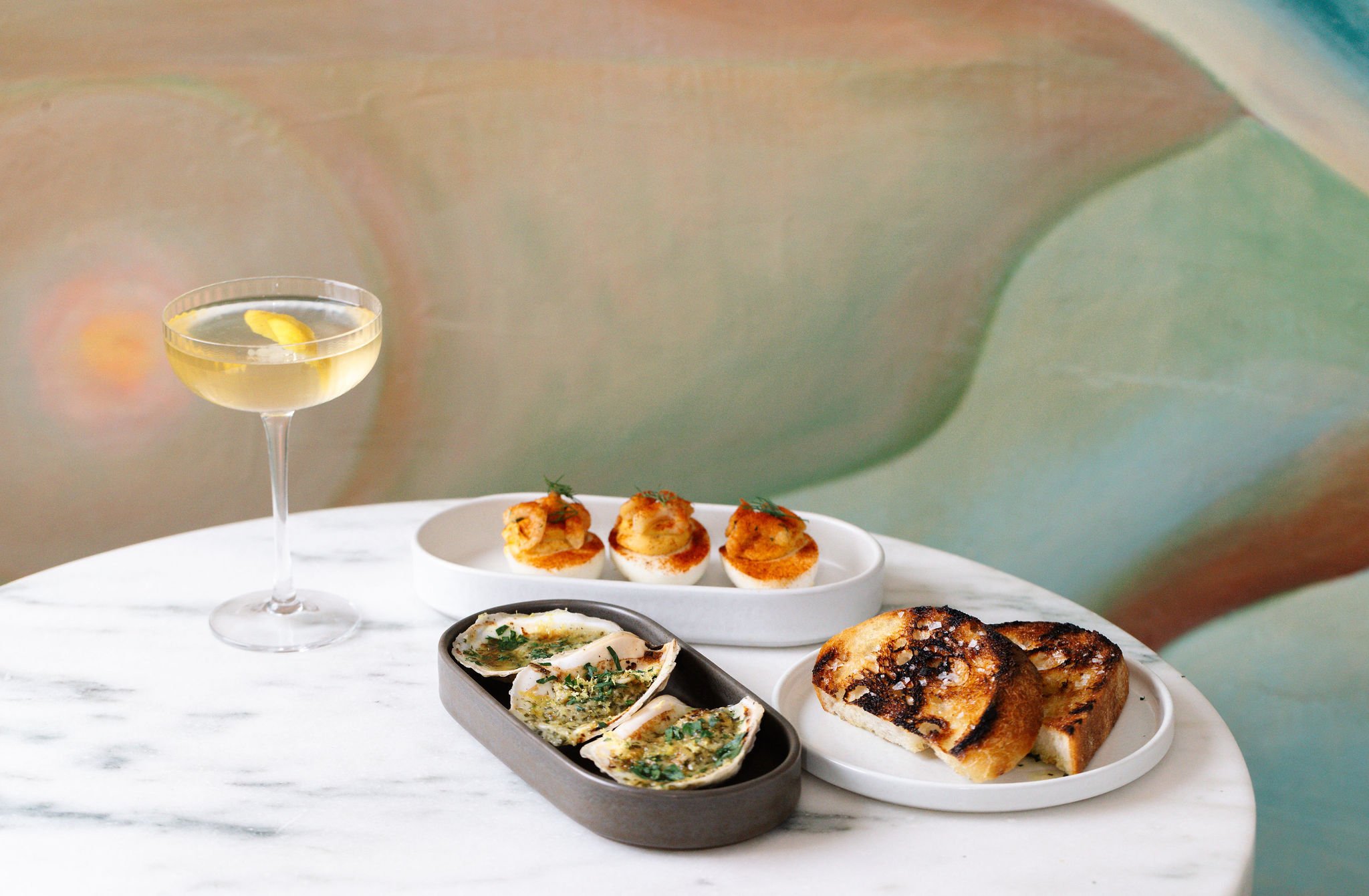 An elegant spread of craft cocktails and small plates at Shy Shy in Chelsea, featuring a Caesar salad martini, broiled oysters, deviled eggs, and toasted sourdough. A perfect selection for a refined yet approachable dining experience.