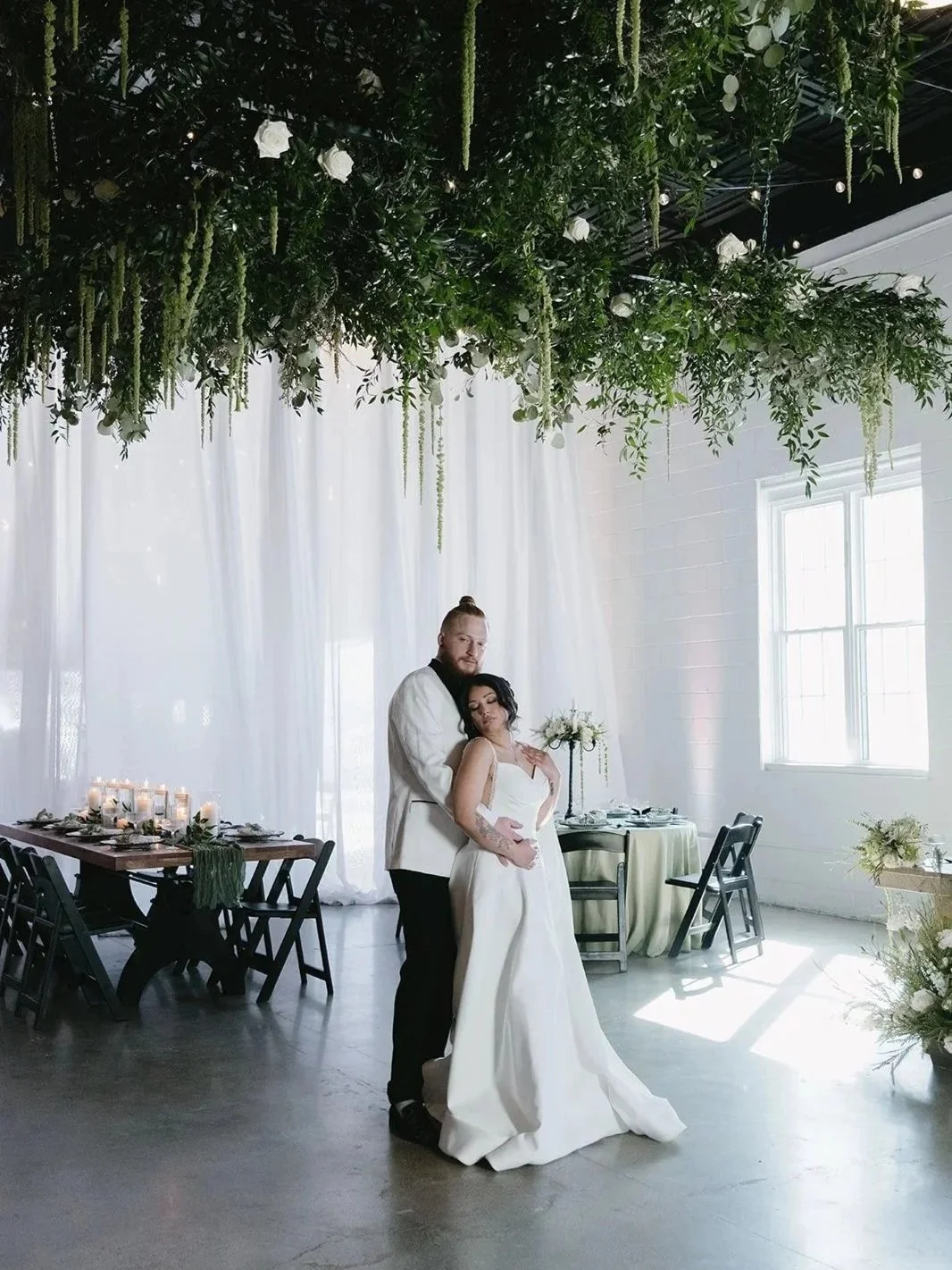a coulpe holds one another under a canopy of lush greens and elegant white florals in a hanging installation at the venues at the factory in Manchester NH