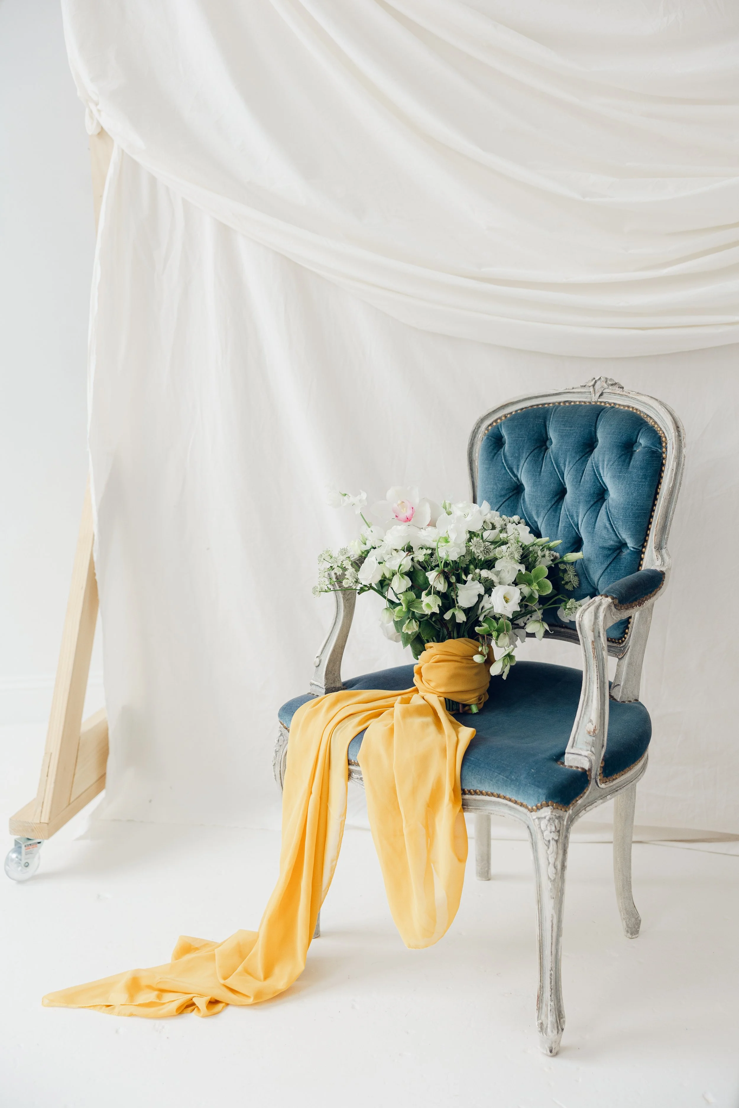 A white artistic bouquet of flowers wrapped in a long mustard yellow fabric fraped over a velvet blue wingback chair inside a bright white studio