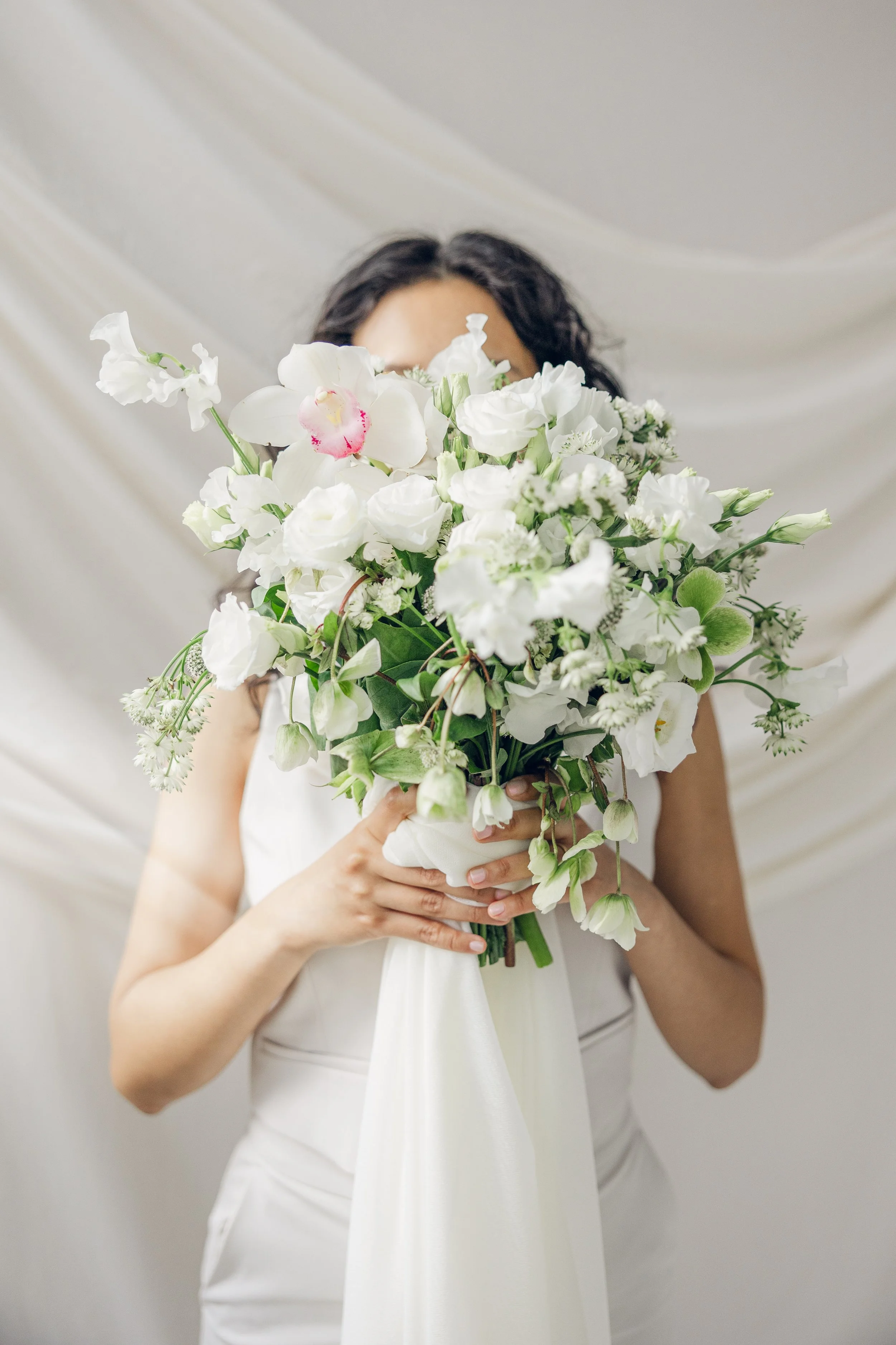 A modern bride holding a elegant white bridal bouquet in front of her face with a white fabric train