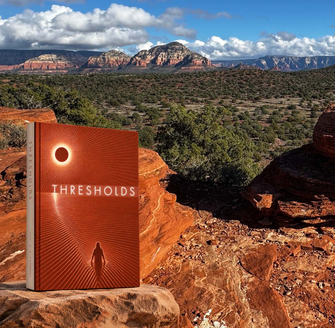A book titled 'Thresholds' with a cover design of an eclipse and a silhouetted figure, placed on a rocky surface in a desert landscape with red rocks, green shrubs, and distant mountains under a partly cloudy sky.