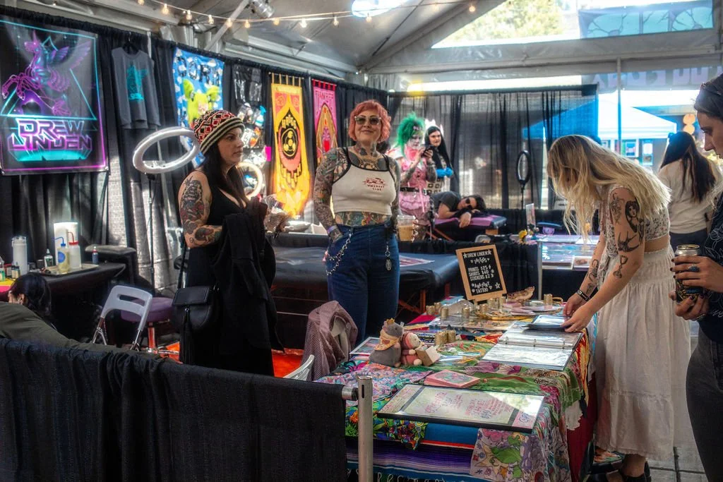 Tattoo artists and customers at a tattoo convention booth with colorful artwork and tattoo designs on display.