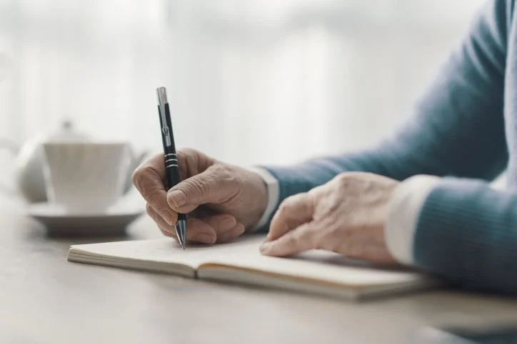 A person writes in a notebook beside a cup.
