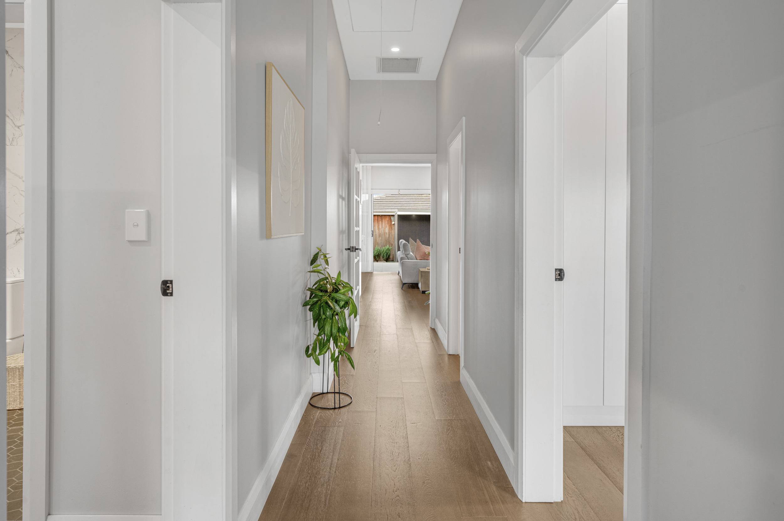 A hallway in a modern home with light gray walls, wooden flooring, white door frames, and a doorway at the end leading to a living room with sofa and outdoor view.