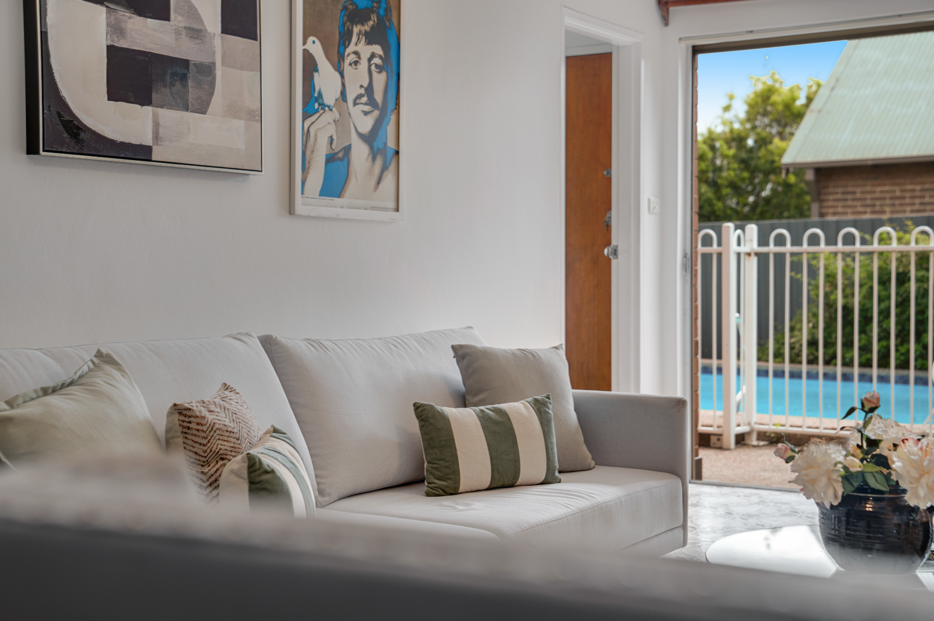 Living room with white couch and decorative pillows, artwork on the wall, open door to backyard with pool, and a bouquet of flowers on a table.