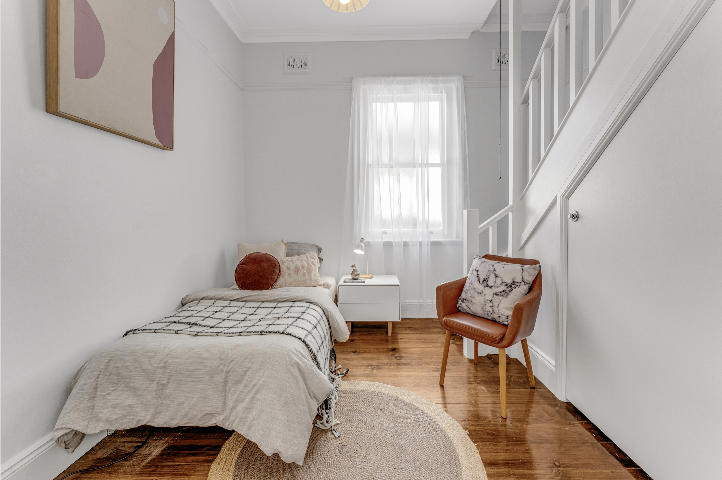 A cozy bedroom with a bed covered in beige and white bedding, a round rust-colored pillow, and a patterned pillow. A white nightstand with a lamp and a small decorative item sits beside the bed. There is a window with sheer white curtains letting in 