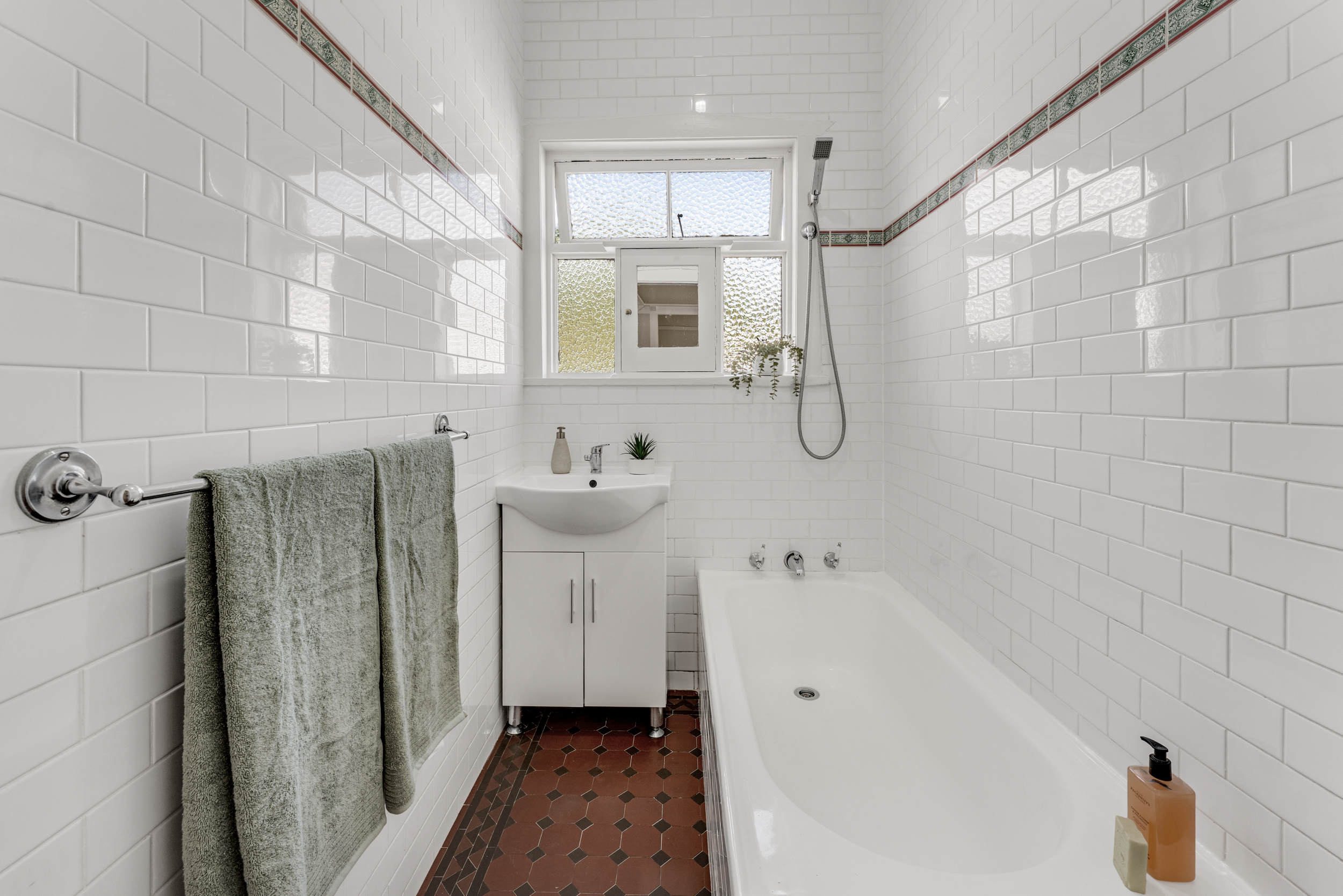 A small bathroom with white tiled walls, a bathtub, a sink with a cabinet, a window with frosted glass, a towel rail with green towels, a plant, and some bottles of soap.