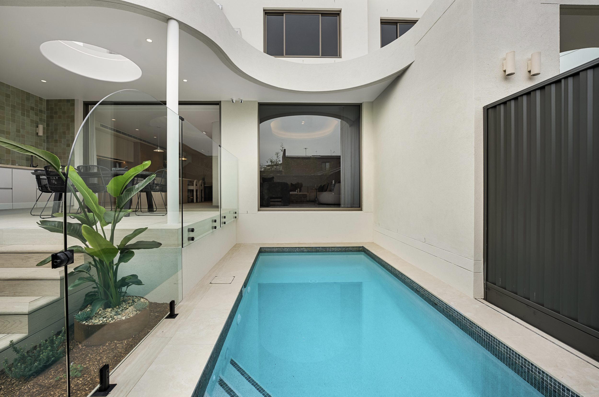 Modern residential building with a small pool on a patio, balcony with curved railing, large windows, and plants in a pot.