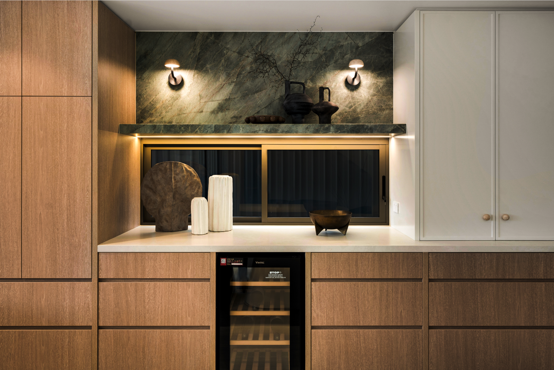 Modern kitchen cabinet with a built-in wine cooler, decorated with artistic vases and sculptures, illuminated by wall-mounted lights and ambient lighting.