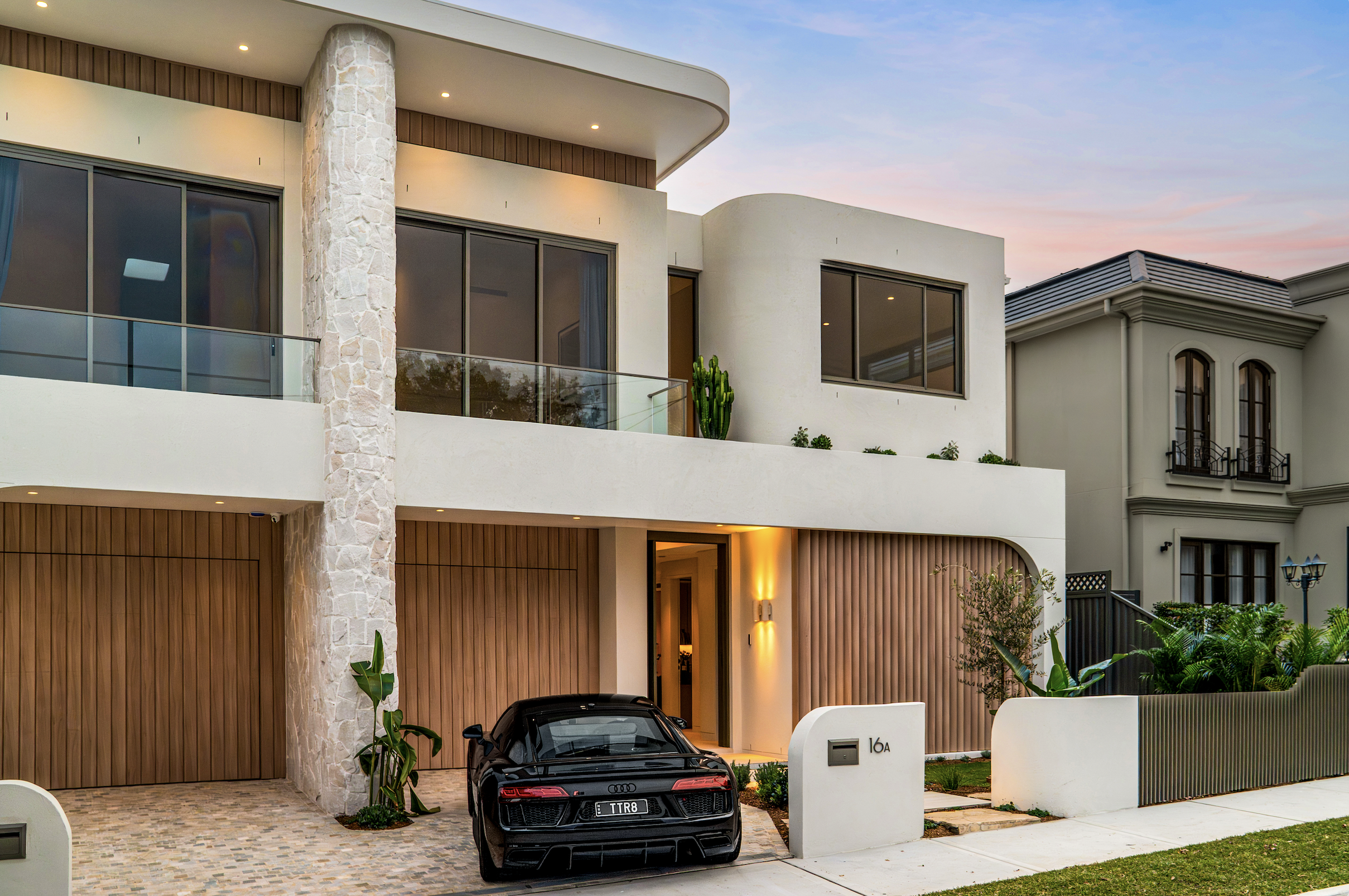 Modern multi-story house with a black sports car parked in front, featuring large windows, a stone column, and a wooden door, with a landscaped yard and a sunset sky in the background.
