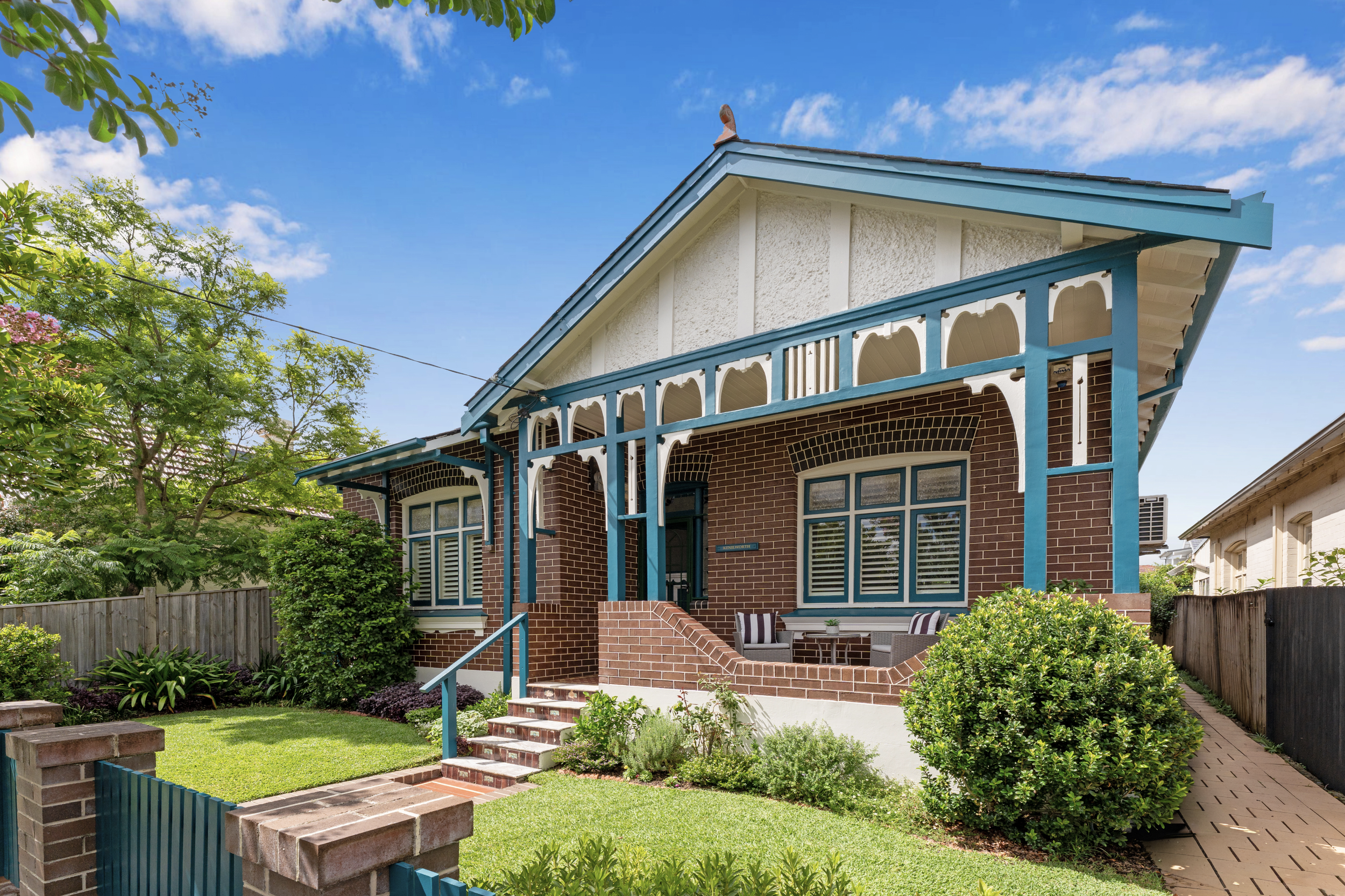 A two-story house with a brick exterior and decorative blue trim, featuring a large front porch with seating, surrounded by greenery and trees under a blue sky with clouds.