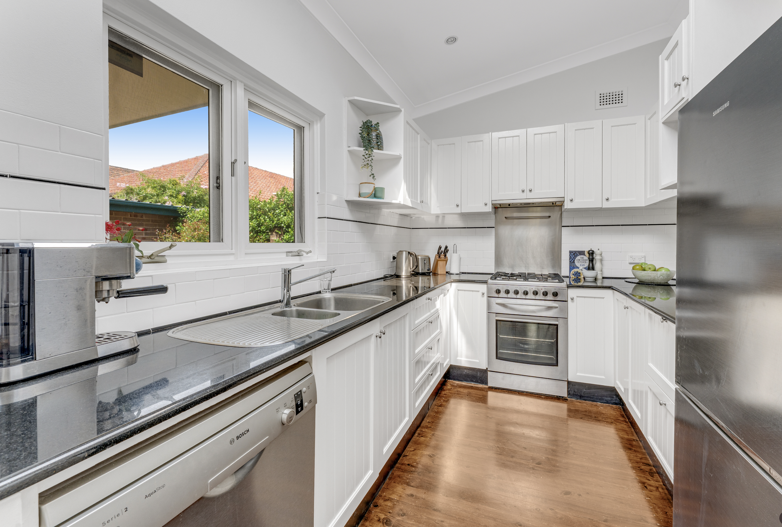 A bright kitchen with white cabinets, stainless steel appliances, and a window showing a view of trees and rooftops. The kitchen has a black countertop, a dishwasher, a stove, and a bowl of green apples.
