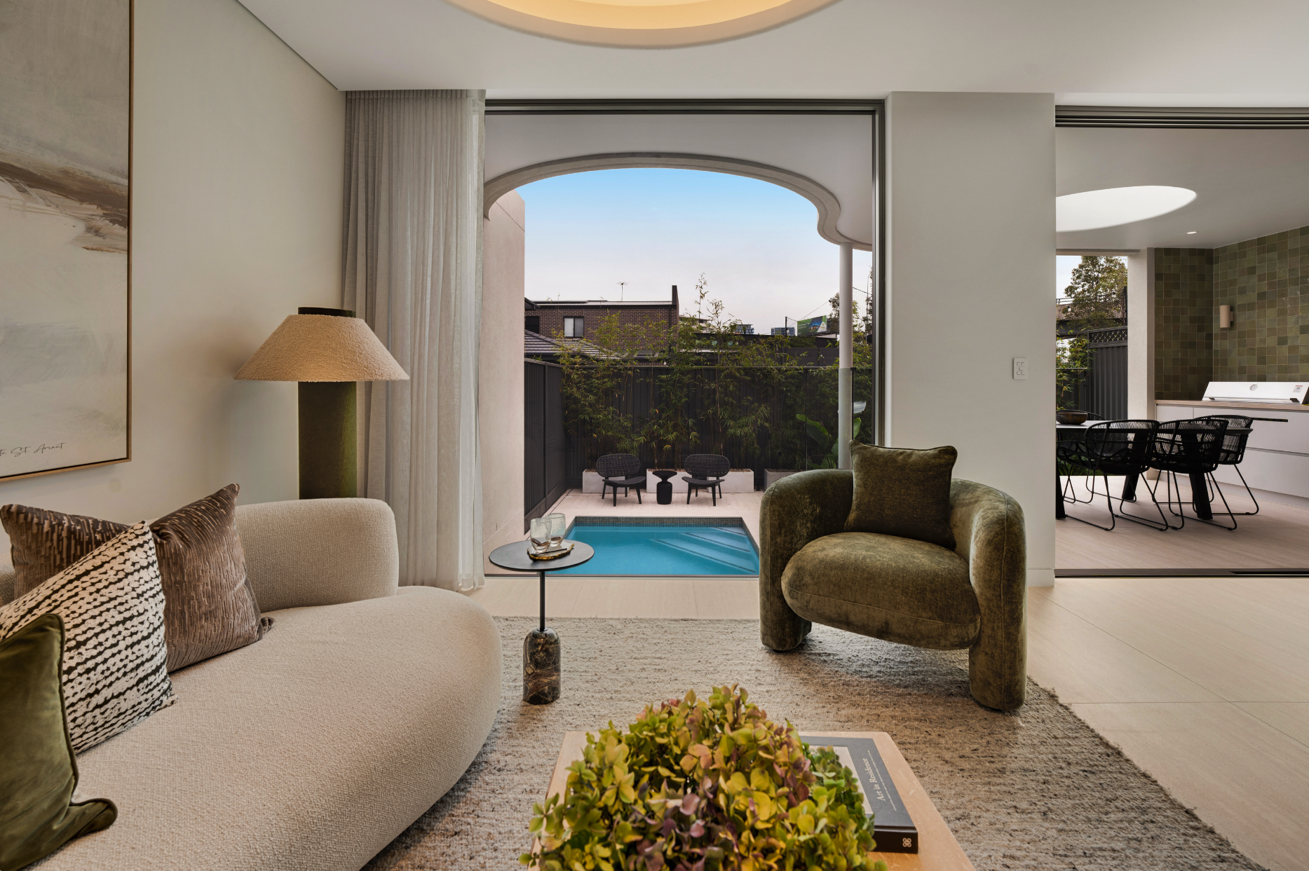 Modern living room with a view of a small patio and pool outside. The room features a beige sofa with patterned and textured pillows, a small round side table with glasses, and a green velvet armchair. There is a floor lamp with a beige shade and a l