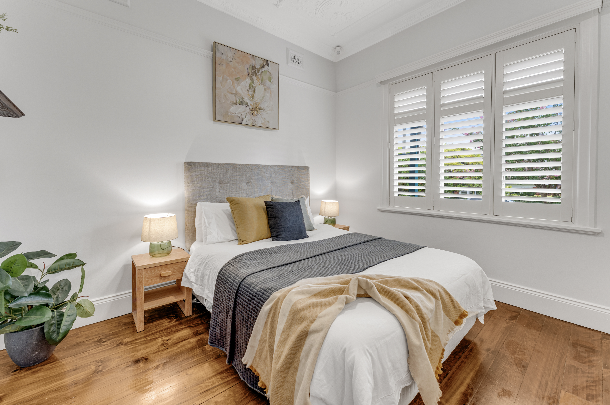 A cozy bedroom with a bed dressed in white, brown, and black bedding, flanked by matching wooden nightstands with lamps. A large window with white shutters and greenery outside. A potted plant in the corner and a floral piece of art on the wall.