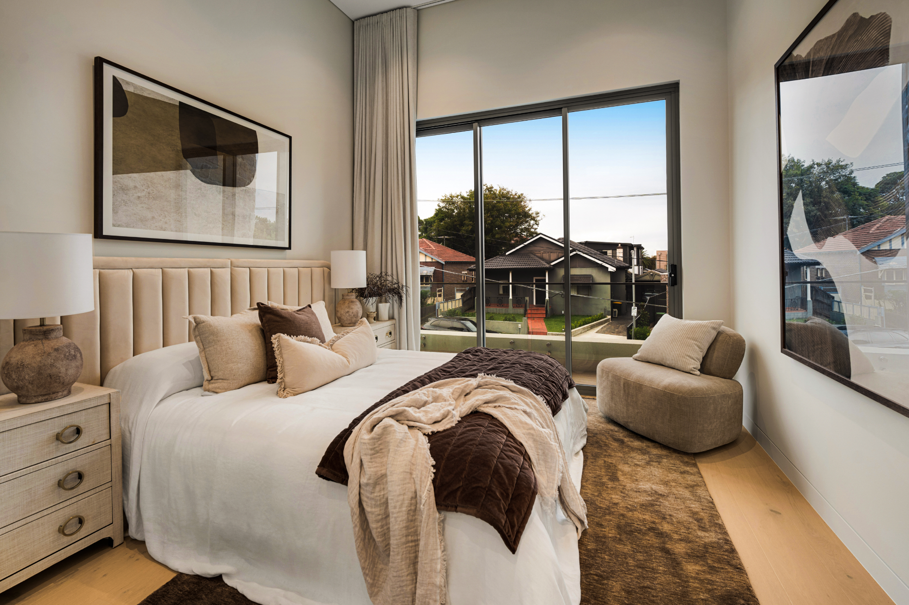 A bedroom with a large window showing a suburban neighborhood. It has a beige upholstered headboard, a nightstand with two lamps, and multiple pillows on the bed with brown and cream bedding. There is an abstract art piece above the bed and another large reflective artwork on the wall opposite the window. A small armchair is placed near the window.