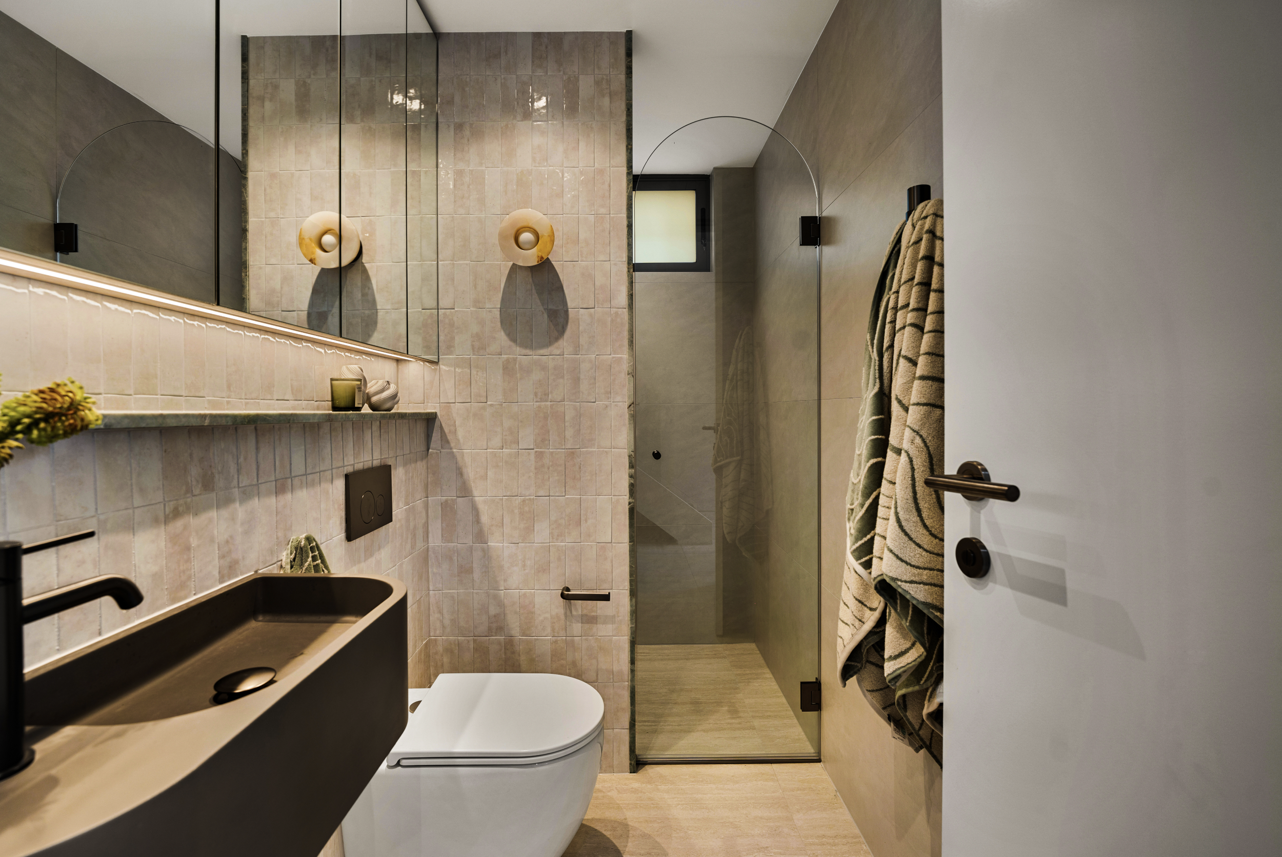 Modern bathroom with beige tiles, a mirror, wall-mounted light fixtures, a black sink, a white toilet, a glass shower with a frosted window, and towels hanging on the wall.