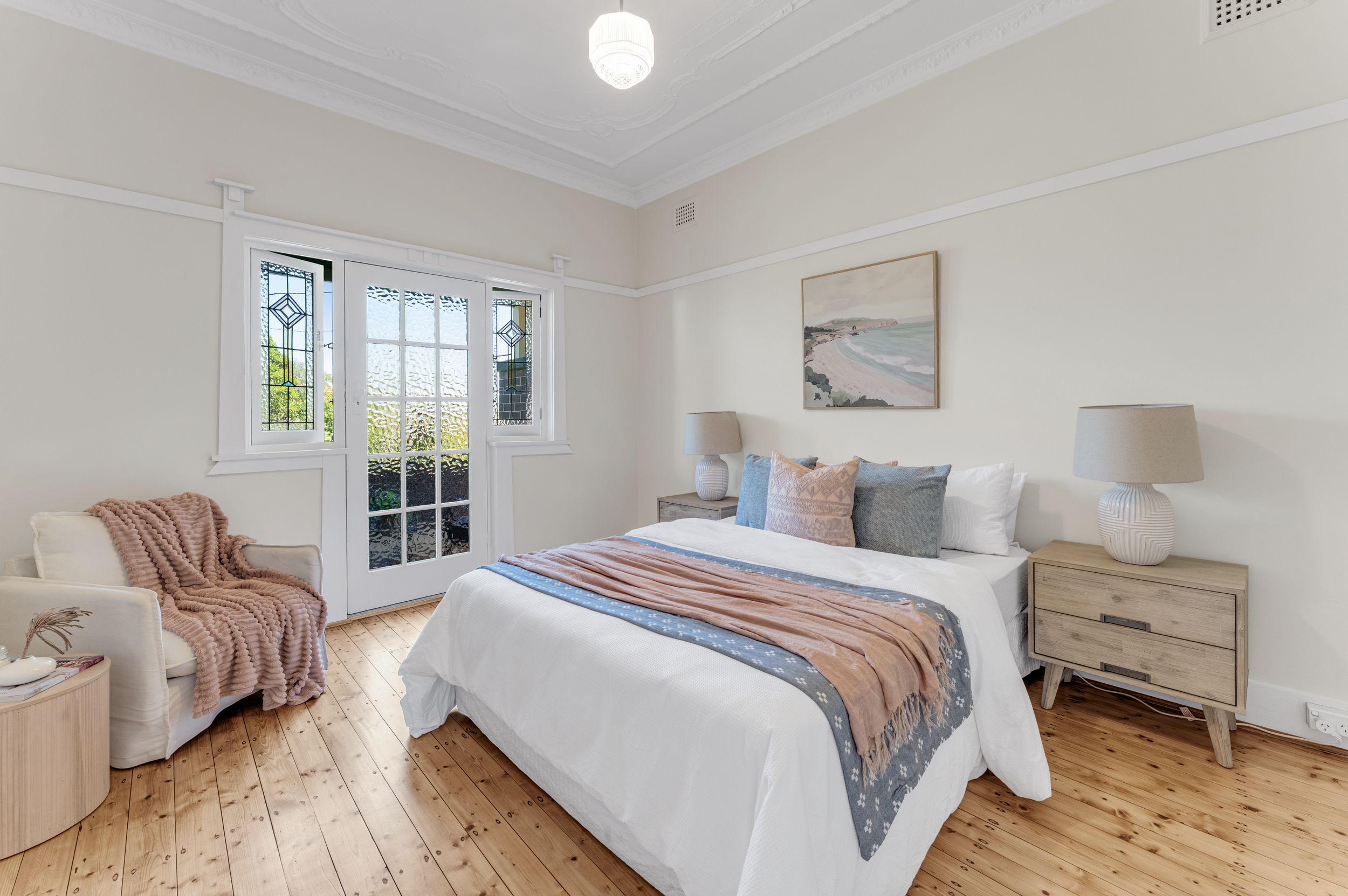 A bedroom with a white bed, wooden nightstands, and matching lamps. There are framed art pieces on the wall, a window with stained glass panels, and a beige armchair with a pink textured throw blanket.