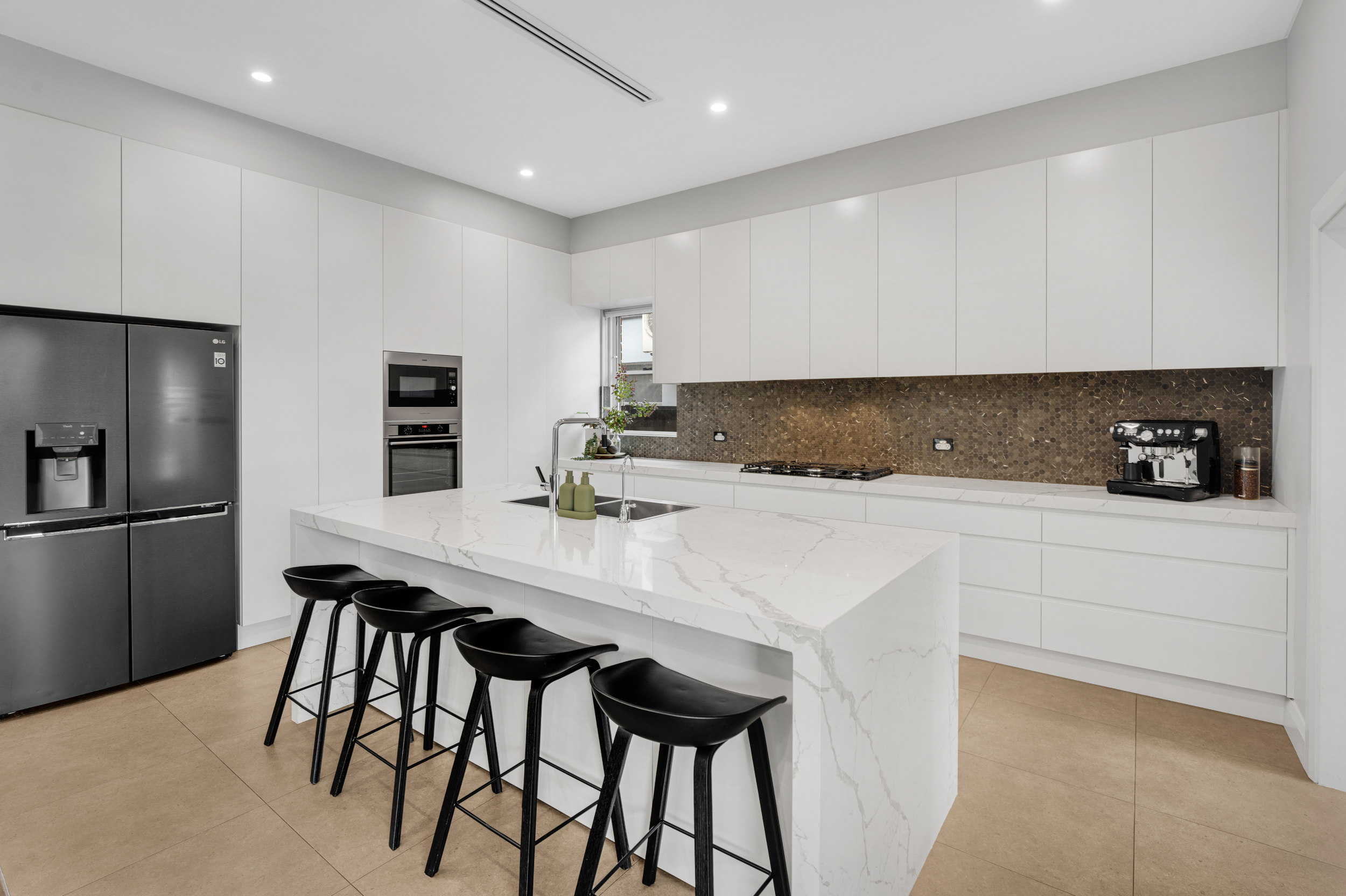 Modern kitchen with white cabinets, marble island, stainless steel refrigerator, black bar stools, and brown mosaic backsplash.