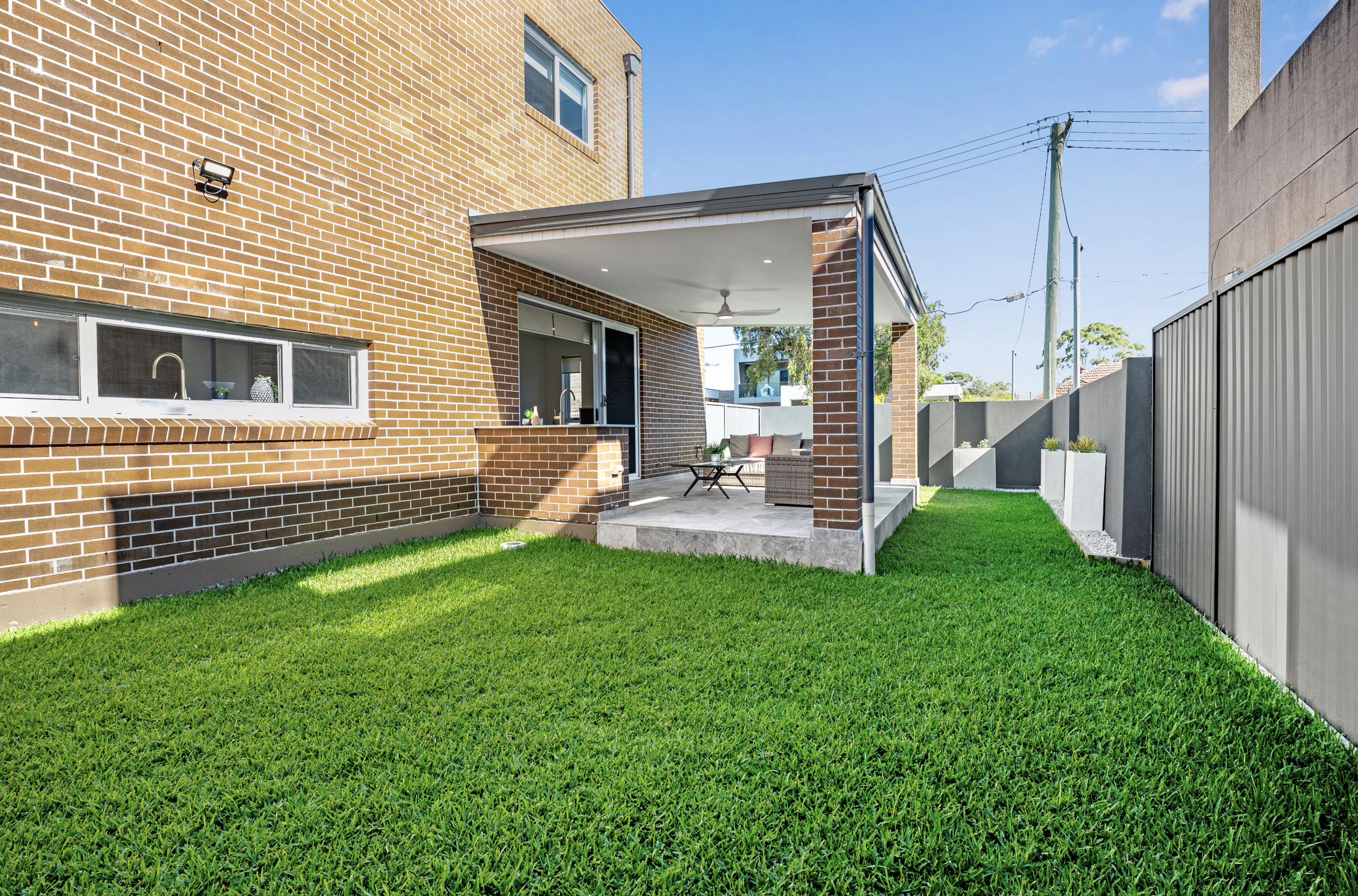 A backyard with green grass, a brick house with an outdoor patio, and a gray fence under a clear blue sky.