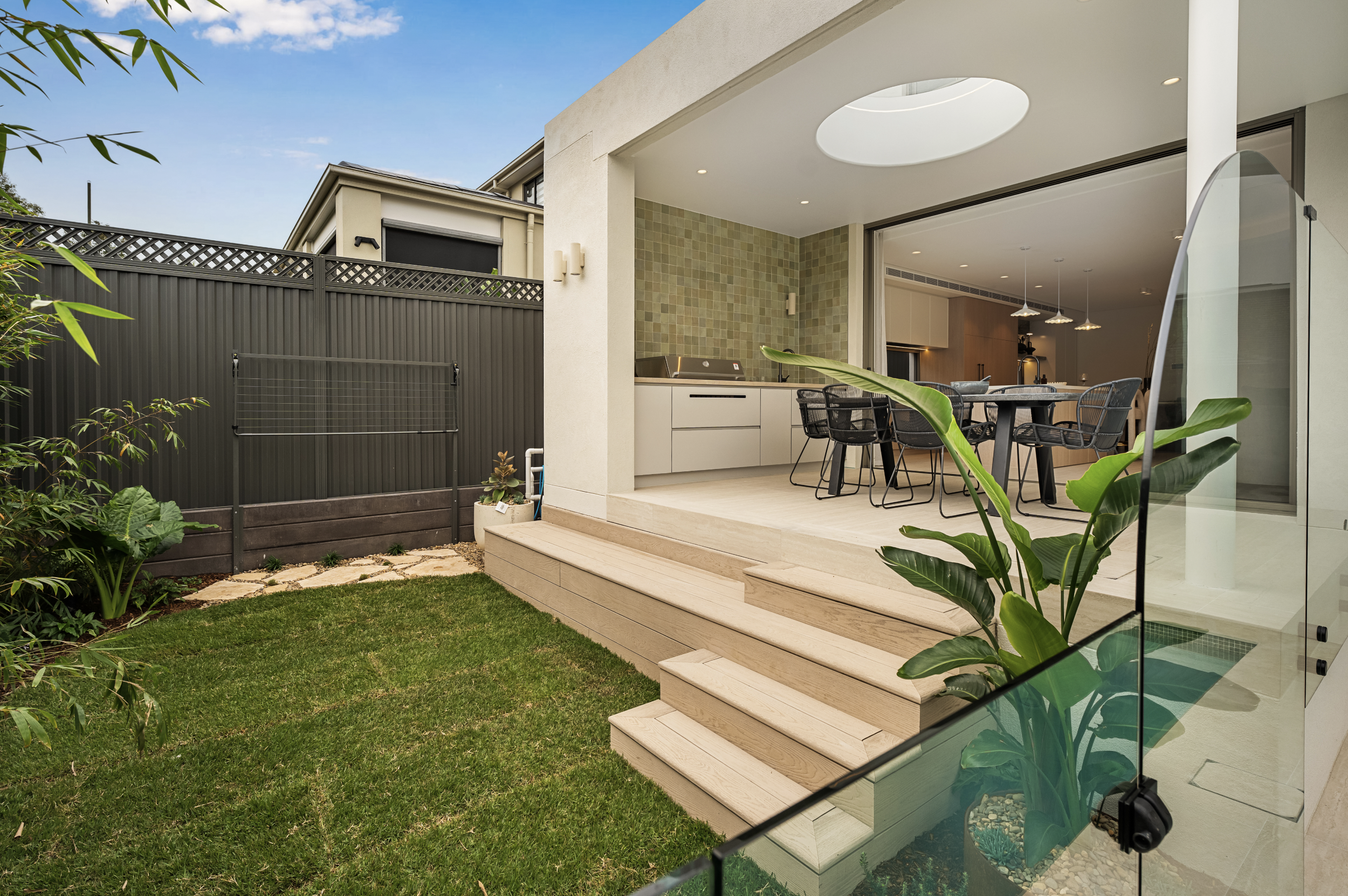Modern backyard patio with steps leading up to a built-in outdoor kitchen and dining area, enclosed by a glass fence, with green grass and plants in the foreground.