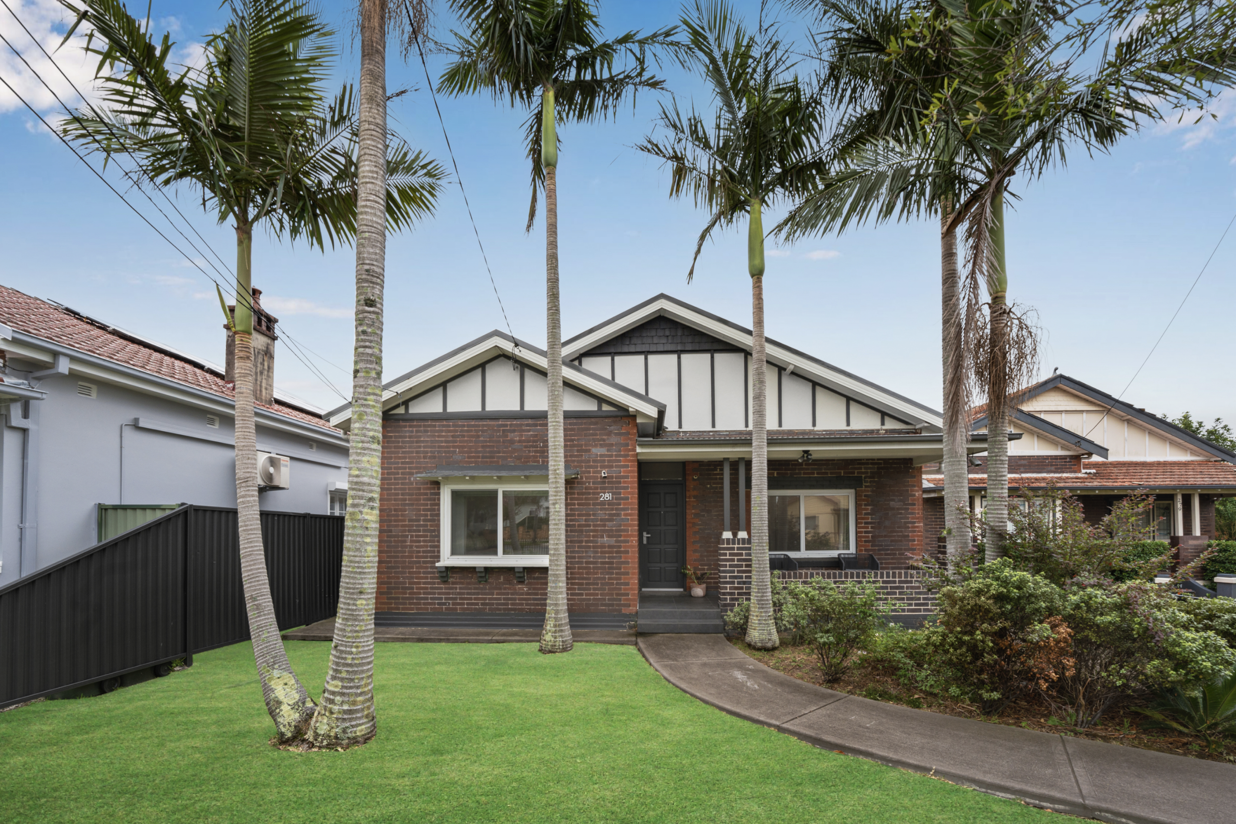 Front view of a house with a brick exterior, black front door, and white window frames, surrounded by palm trees and a well-maintained lawn.