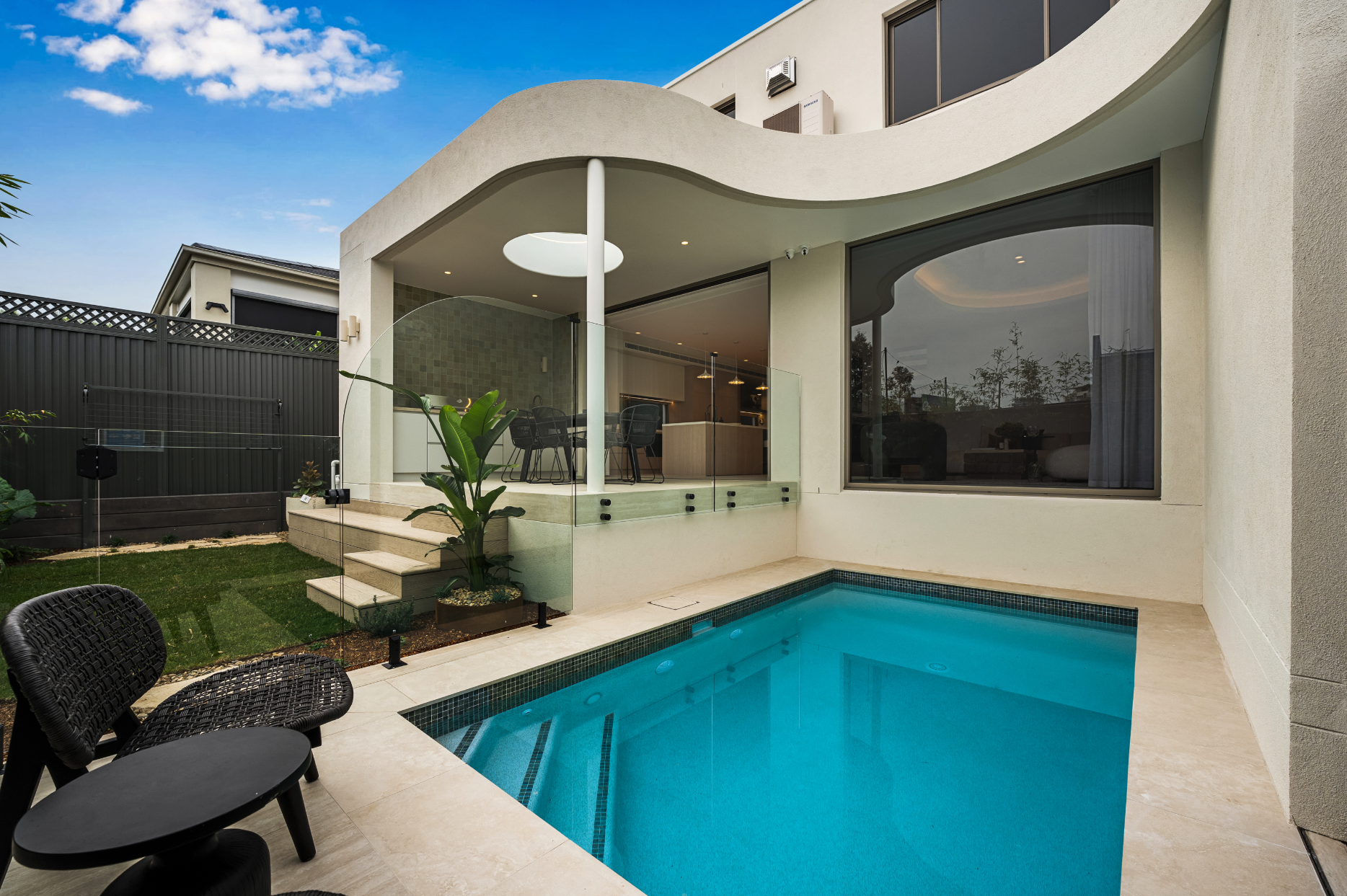 Modern backyard with a swimming pool, outdoor seating, and a glass patio enclosed with a view of the house interior through large windows.