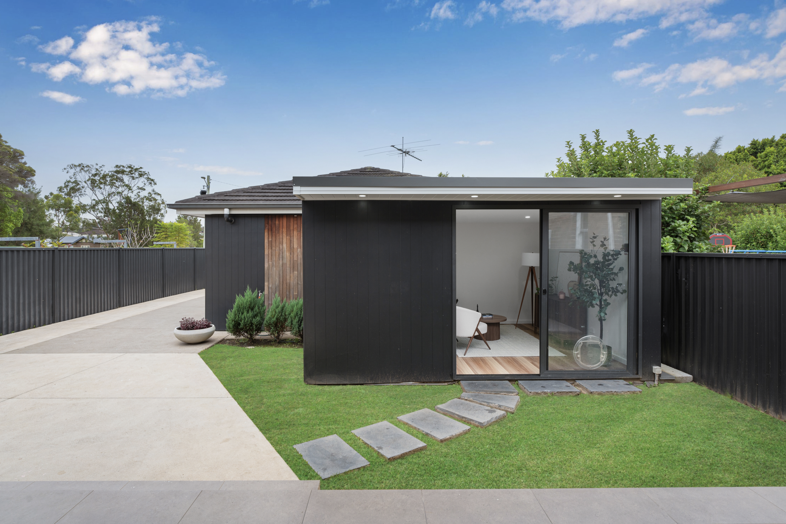 A modern house with dark exterior walls and a sliding glass door leading inside. A small yard with green grass, potted plants, and a pathway of stepping stones. The sky is blue with some scattered clouds.