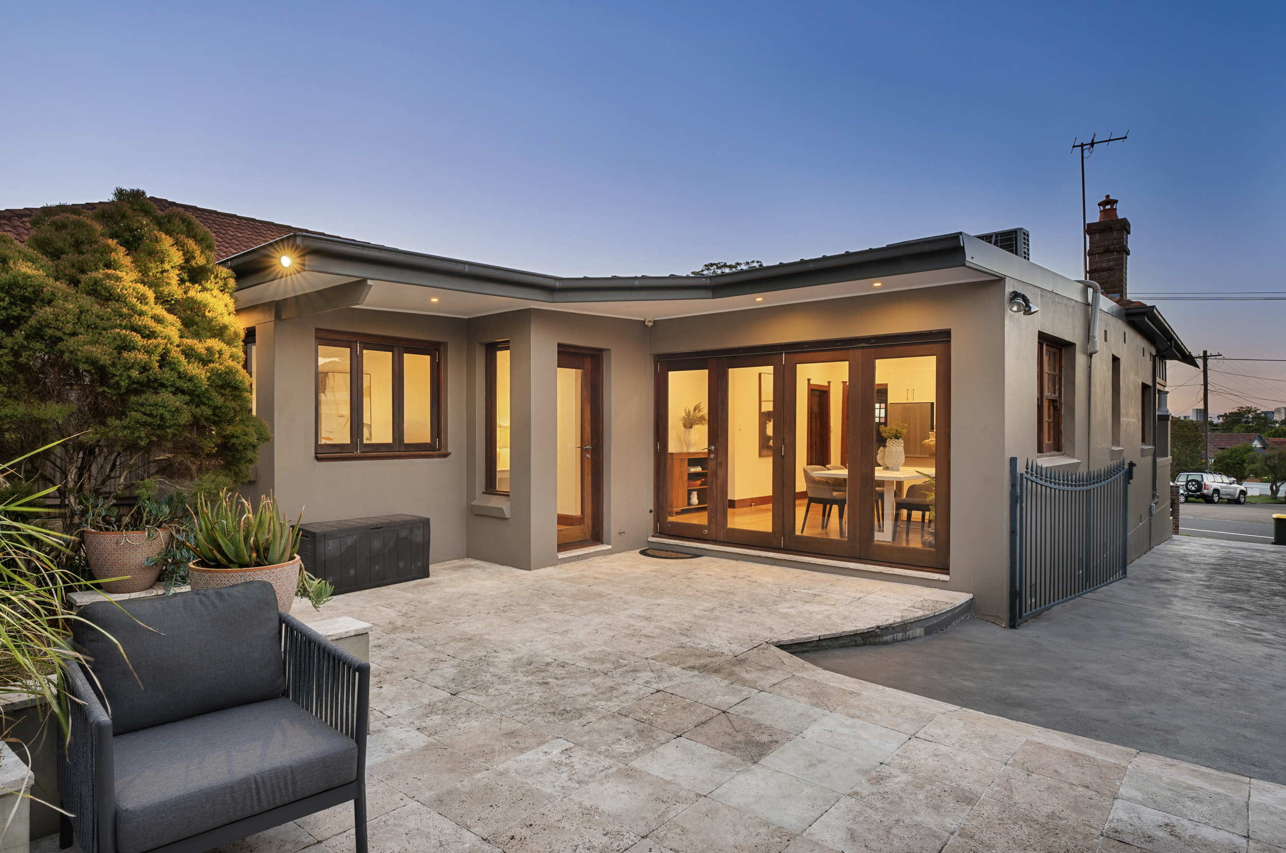 Backyard patio area of a house during evening with outdoor furniture, potted plants, and large windows with warm interior lighting.