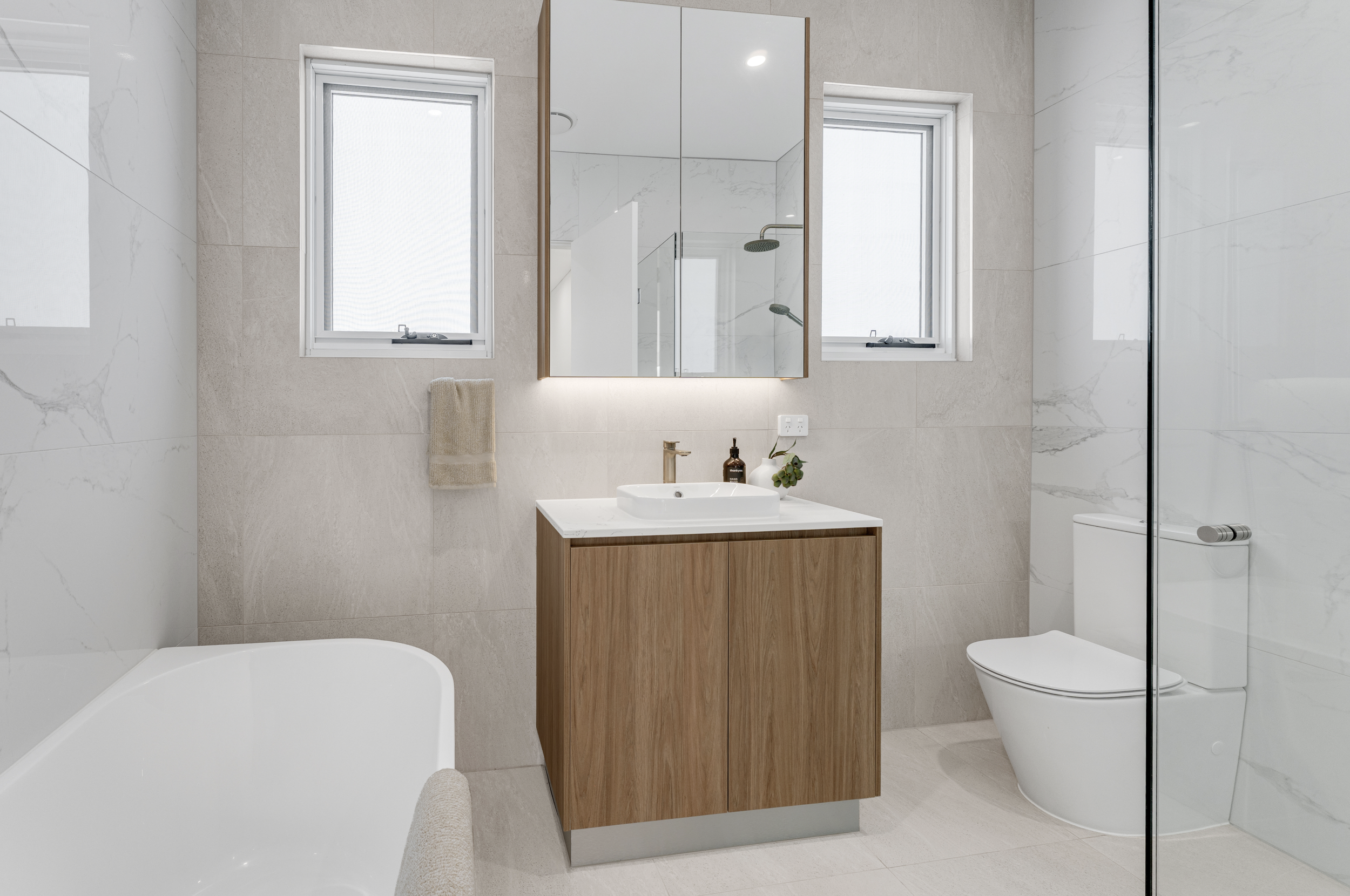 Modern bathroom with beige tiled walls, two windows, a mirror cabinet, a white sink with a wooden cabinet, a bottle of soap, a small plant, a towel, a toilet, and a walk-in shower with a glass partition.
