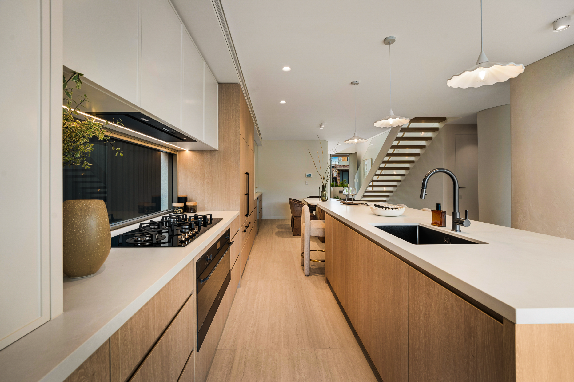 A modern kitchen with light wood cabinetry, a white countertop island, black sink, and pendant lights. There is a staircase with open risers in the background and small dining area with chairs and decorative vases.