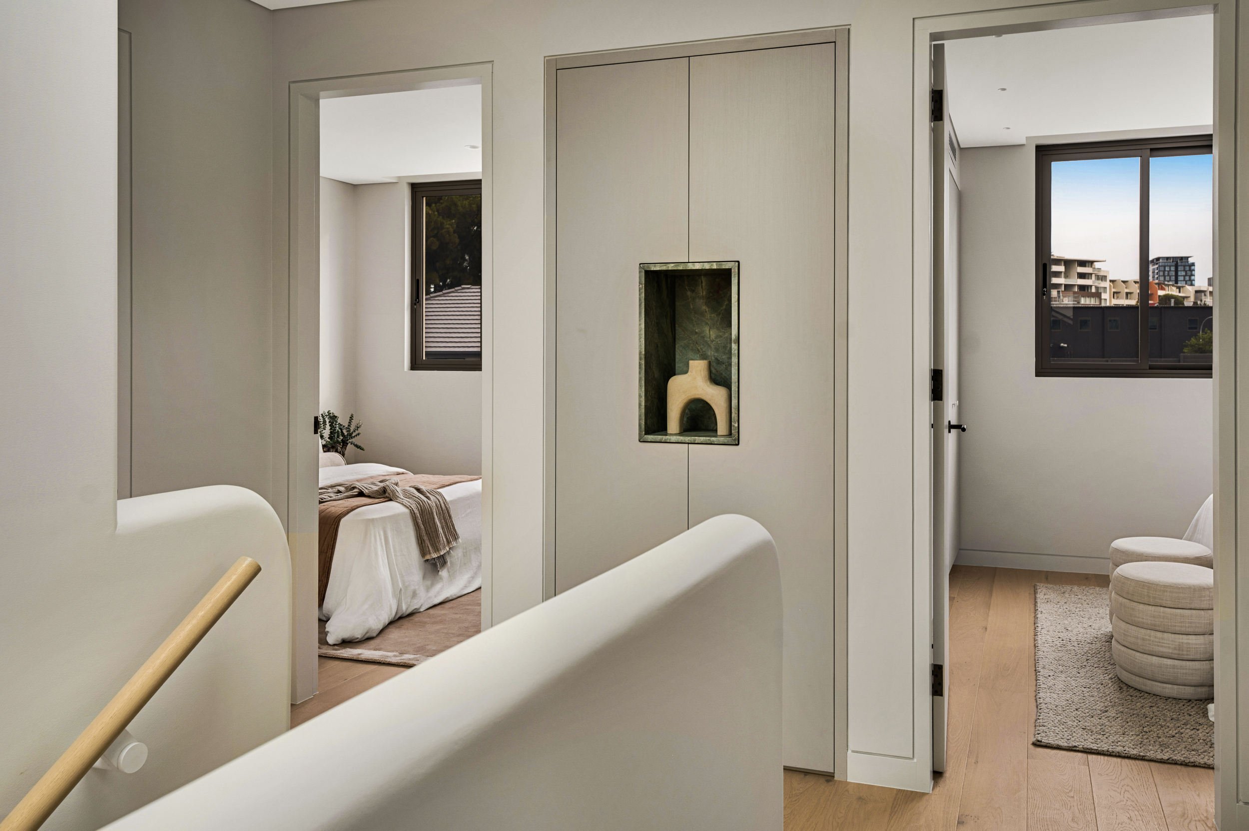 A view of a bedroom with large windows and a modern interior, featuring a bed with neutral bedding and a decorative shelf with a vase, as seen from the perspective of a hallway or adjacent room.