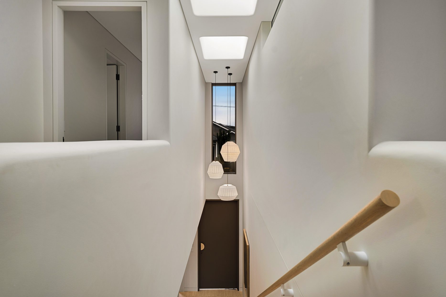 Interior view of a modern stairwell with hanging pendant lights, a window showcasing a blue sky, white walls, and a wooden handrail.