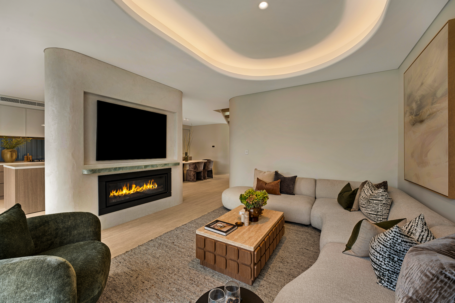 Modern living room with a curved sectional sofa, decorative pillows, a wooden coffee table with a flower arrangement, a built-in fireplace, a large mounted flat-screen TV, and abstract art on the wall. Created with warm lighting and neutral tones.