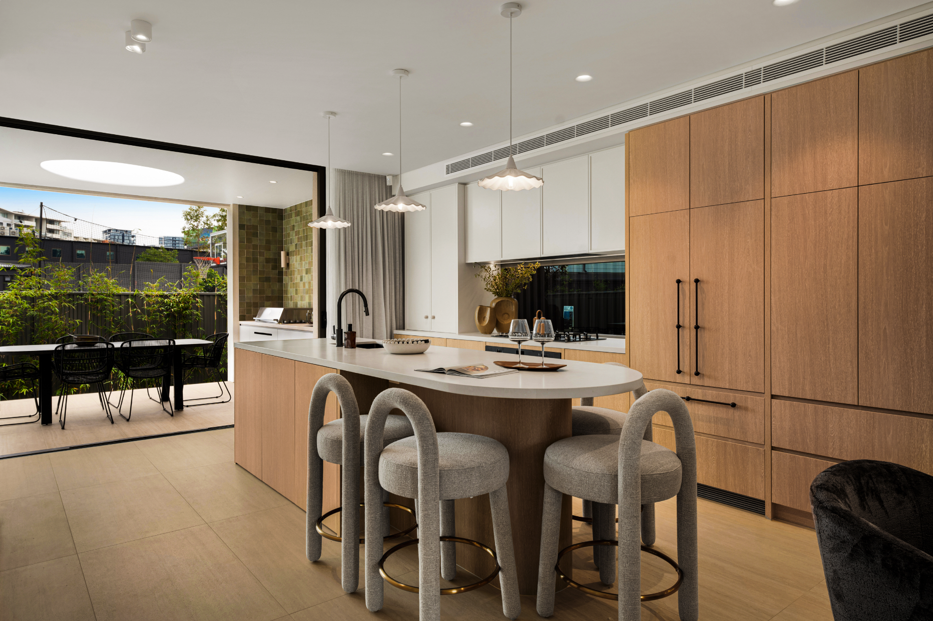 Modern open-concept kitchen and dining area with a large island, light wood cabinetry, and seating for four. There are hanging pendant lights above the island, a built-in oven, and a view of an outdoor patio with a table, chairs, and greenery.